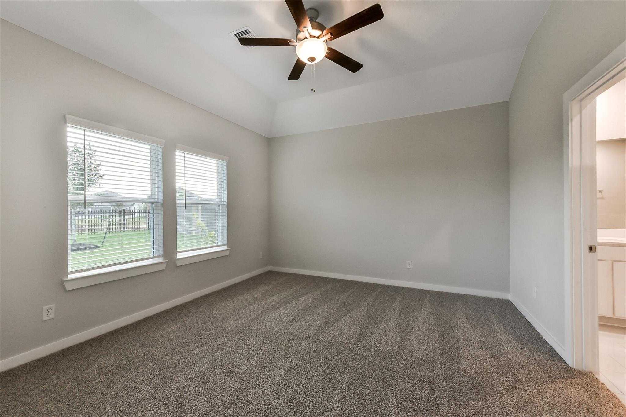 Bright secondary bedroom with beige walls, gray carpet, ceiling fan, large windows, and en-suite bath in Davidson Homes The Laguna B, Sunterra Katy
