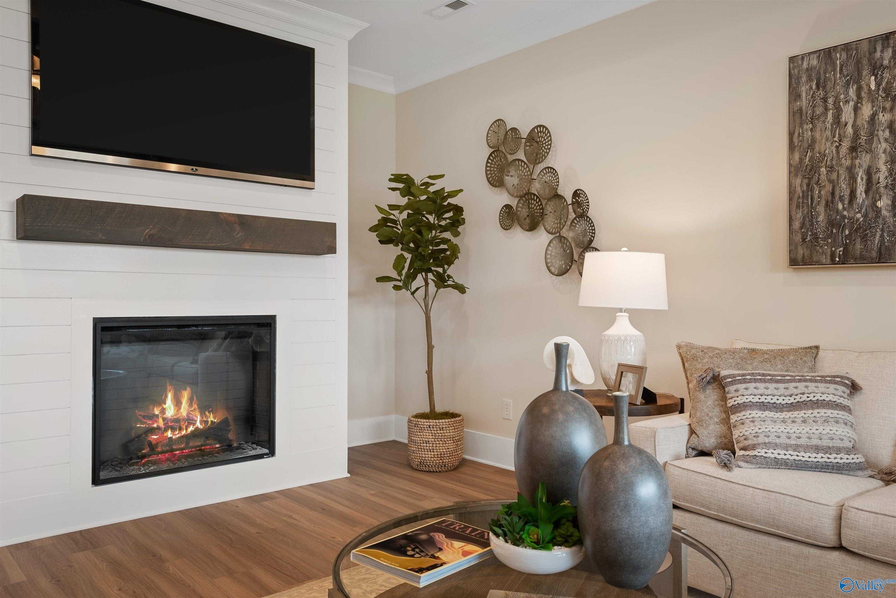 Cozy living room with gas fireplace, wooden mantel, mounted TV, beige sofa, potted plants in Davidson Homes The Everett, New Market, Alabama