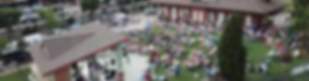 Aerial view of vibrant community event at Sanford pavilion with stage, seated crowd on green lawn, and historic red brick depot amid urban surroundings