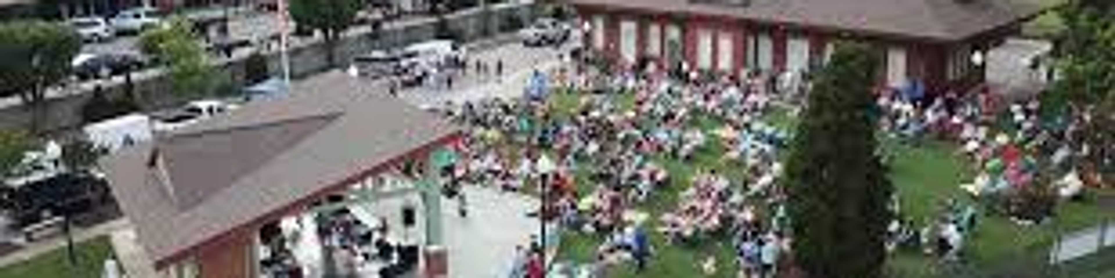 Aerial view of vibrant community event at Sanford pavilion with stage, seated crowd on green lawn, and historic red brick depot amid urban surroundings