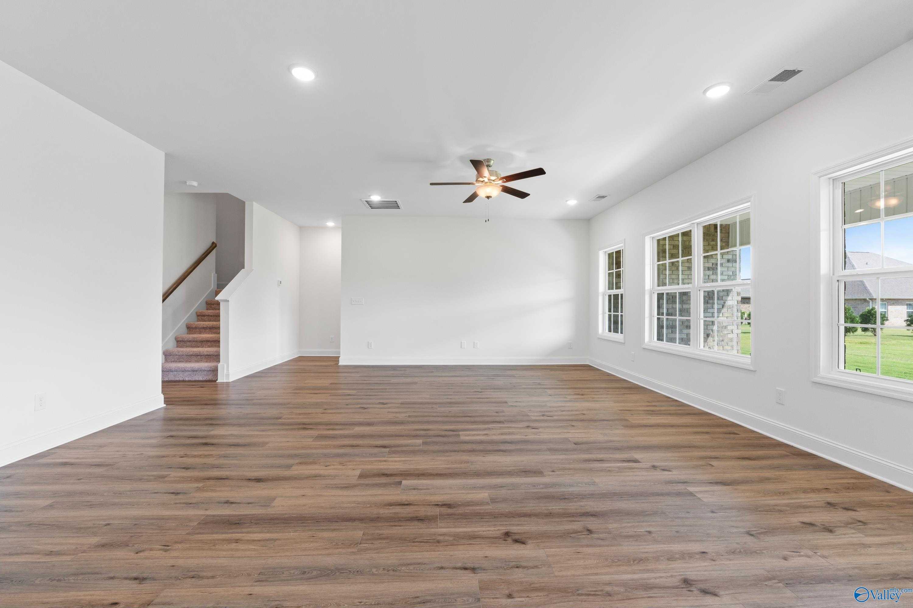 Spacious great room with hardwood floors, dual ceiling fans, large windows, and staircase in Davidson Homes The Rockford B with Bonus, Toney, AL