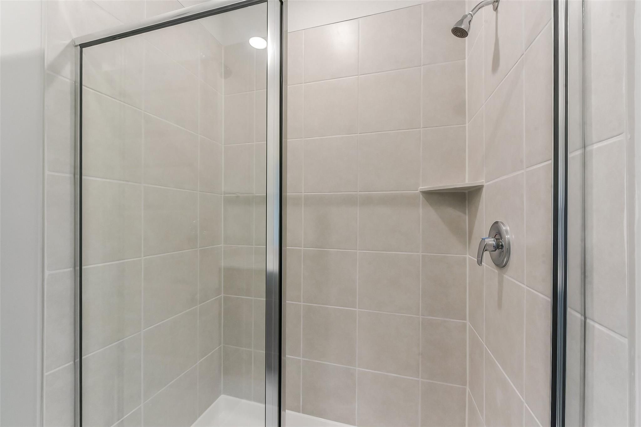 Clear glass shower enclosure with beige subway tiles, niche shelf, and chrome showerhead in master bath of Davidson Homes The Sabine F, Dayton, Texas