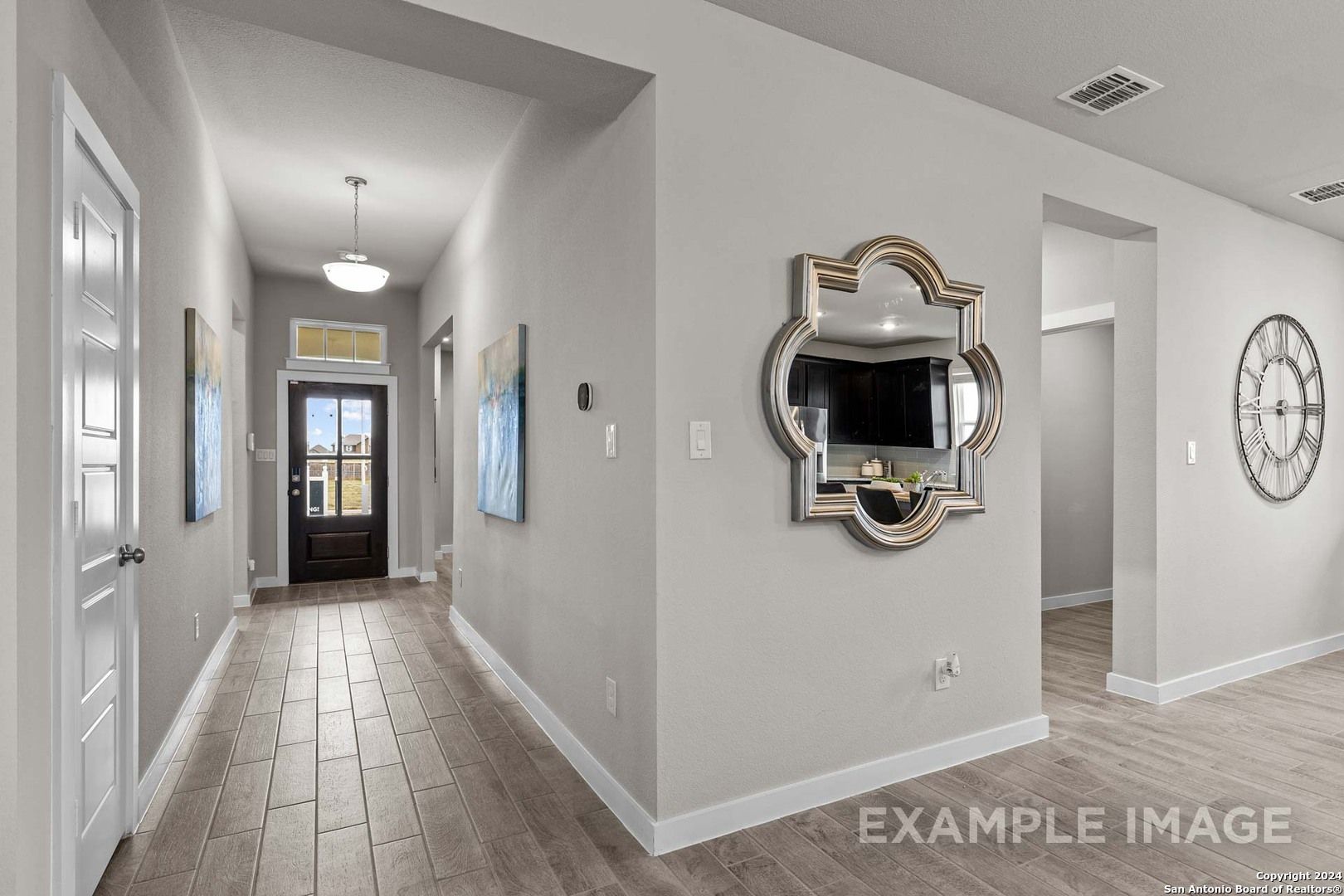 Elegant entry hallway with light wood floors, gray walls, framed mirror TV, wall clock, and front door in Davidson Homes The Collin A, Seguin, Texas