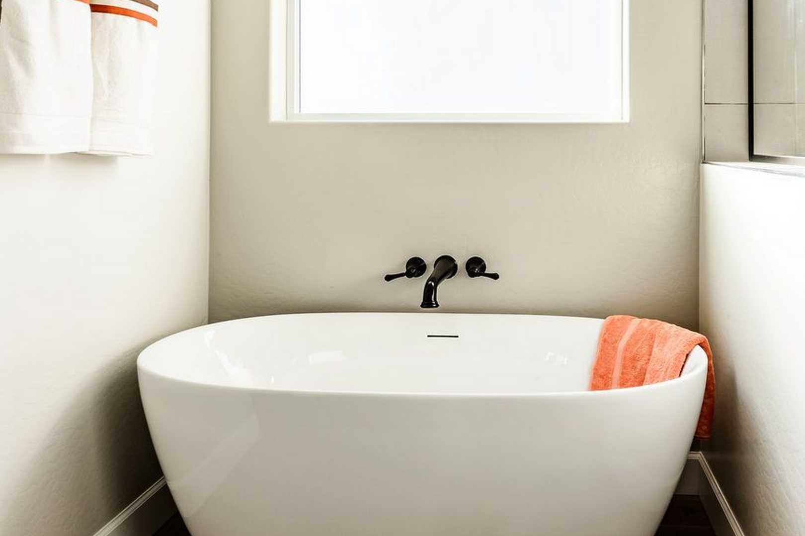 Freestanding white soaking tub in The Monarch master bathroom, black faucet, neutral walls, window