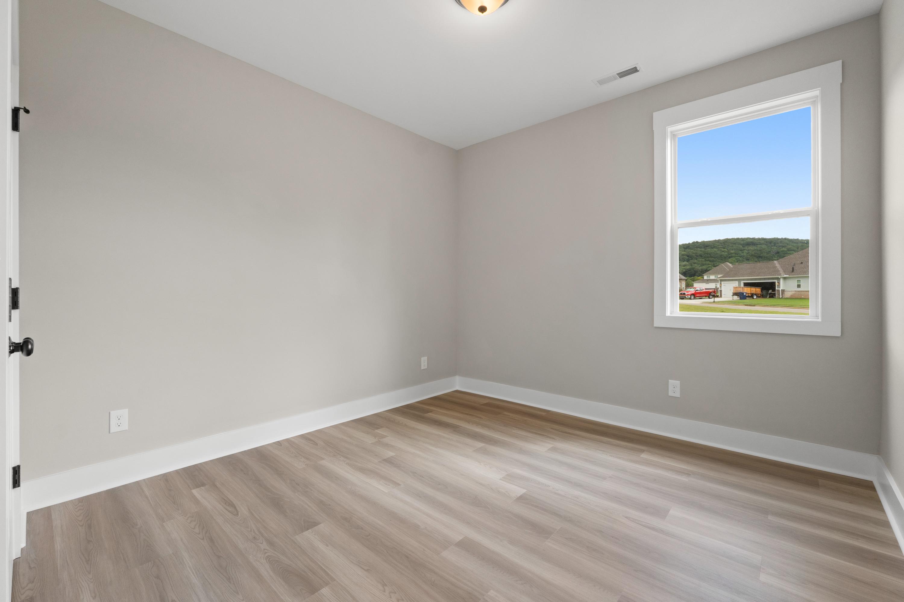 Spacious secondary bedroom in The Oxford B with light gray walls, hardwood floors, and large window overlooking suburban landscape