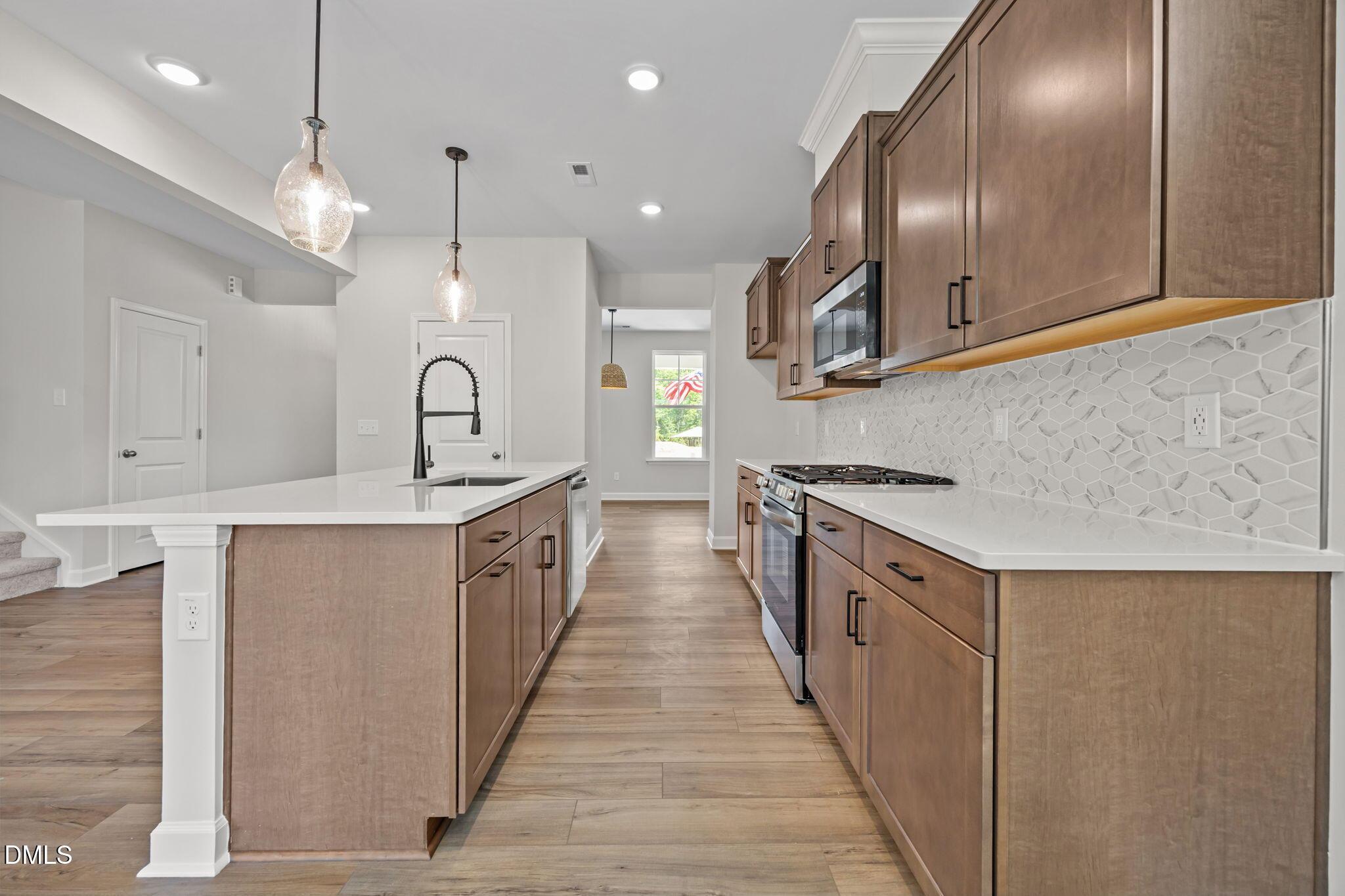 Open-concept kitchen with white quartz island, brown shaker cabinets, stainless appliances, and hexagon tile backsplash in The Willow D, Wendell, NC
