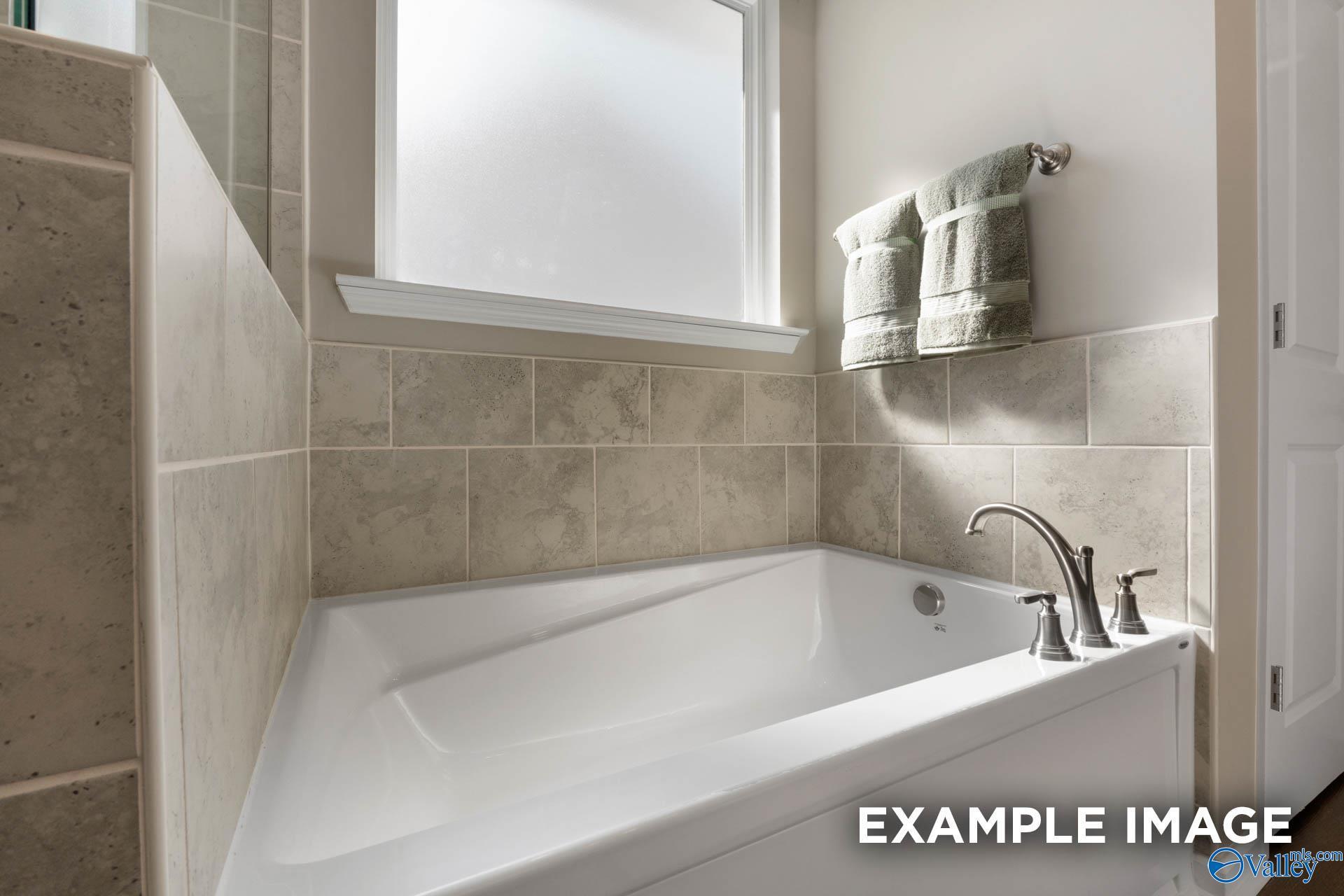Elegant corner soaking tub with beige tile surround and frosted window in The Stella master bath, Madison, Alabama