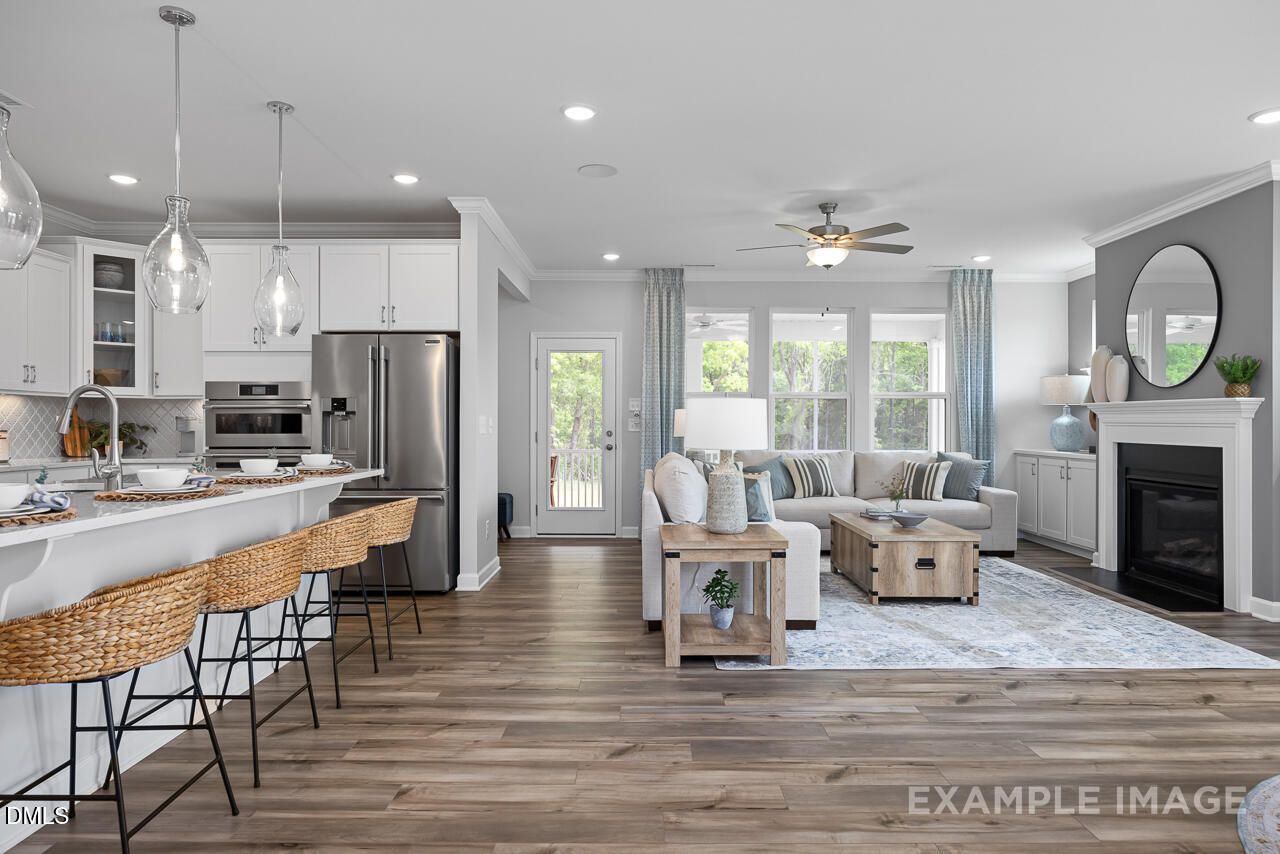 Open-concept living room and kitchen with white cabinets, stainless appliances, island bar stools, gray sectional sofa, and fireplace in Davidson Homes The Magnolia D, Angier, NC
