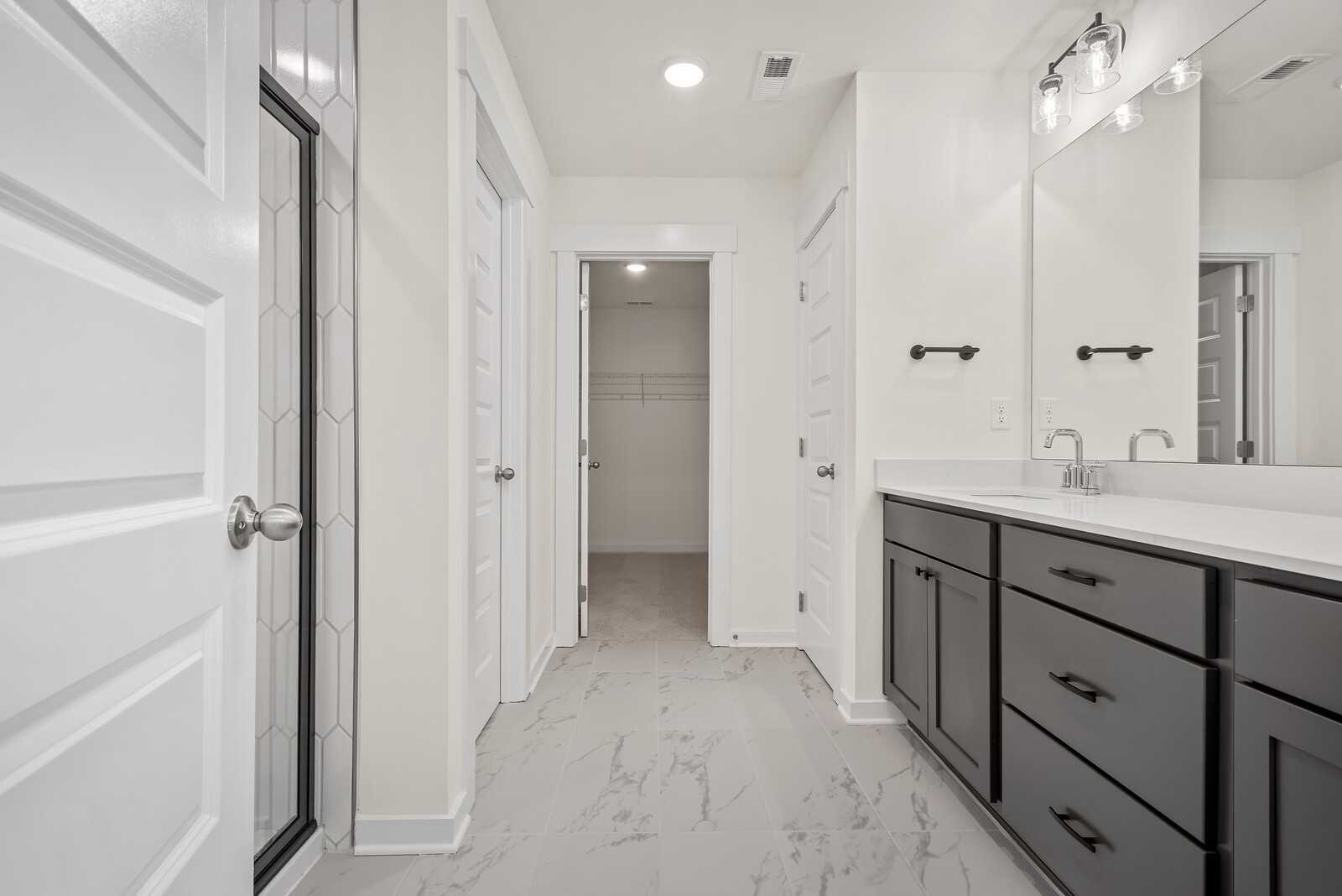 Elegant master bathroom with double dark cabinet vanity, white quartz counters, glass hex-tile shower, and closet access in Davidson Homes The Logan C, Gallatin, TN