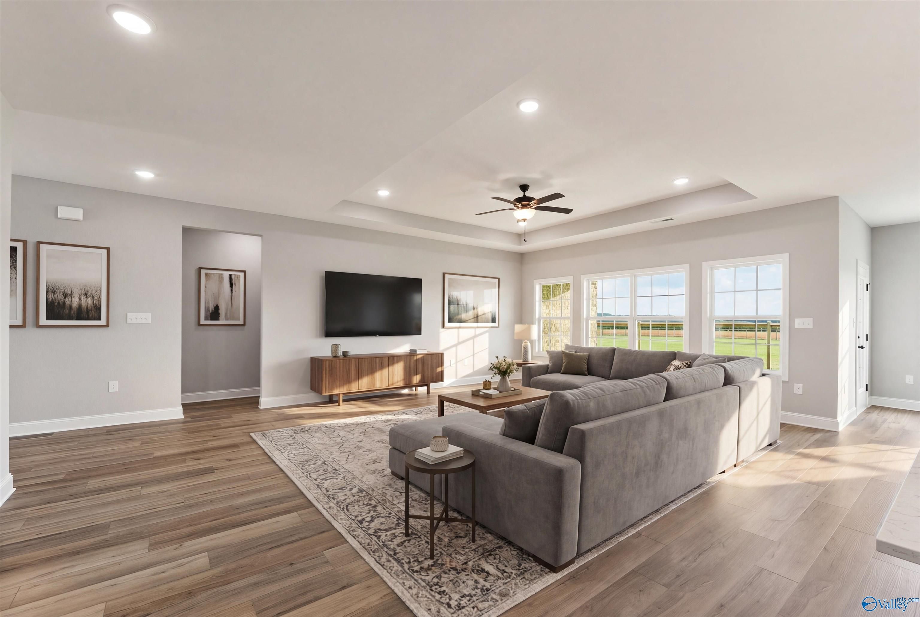Cozy living room with gray L-shaped sectional, ceiling fan, TV wall, hardwood floors, and large windows to fields in Davidson Homes The Finleigh, Toney, Alabama