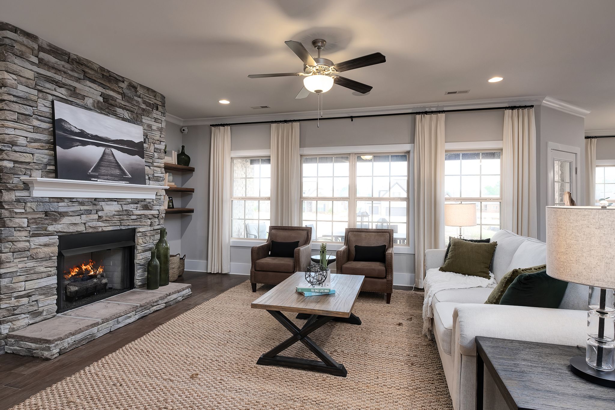 Cozy living room in Chimney Creek at Hampton Cove, Alabama with stacked stone fireplace, burning fire, beige sofa, armchairs, and large curtained windows