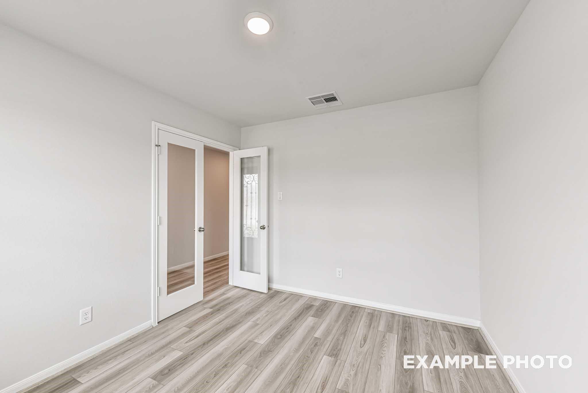 Bright bedroom with open French doors, white walls, and laminate wood floors in Davidson Homes The Riviera A, Rosharon, Texas