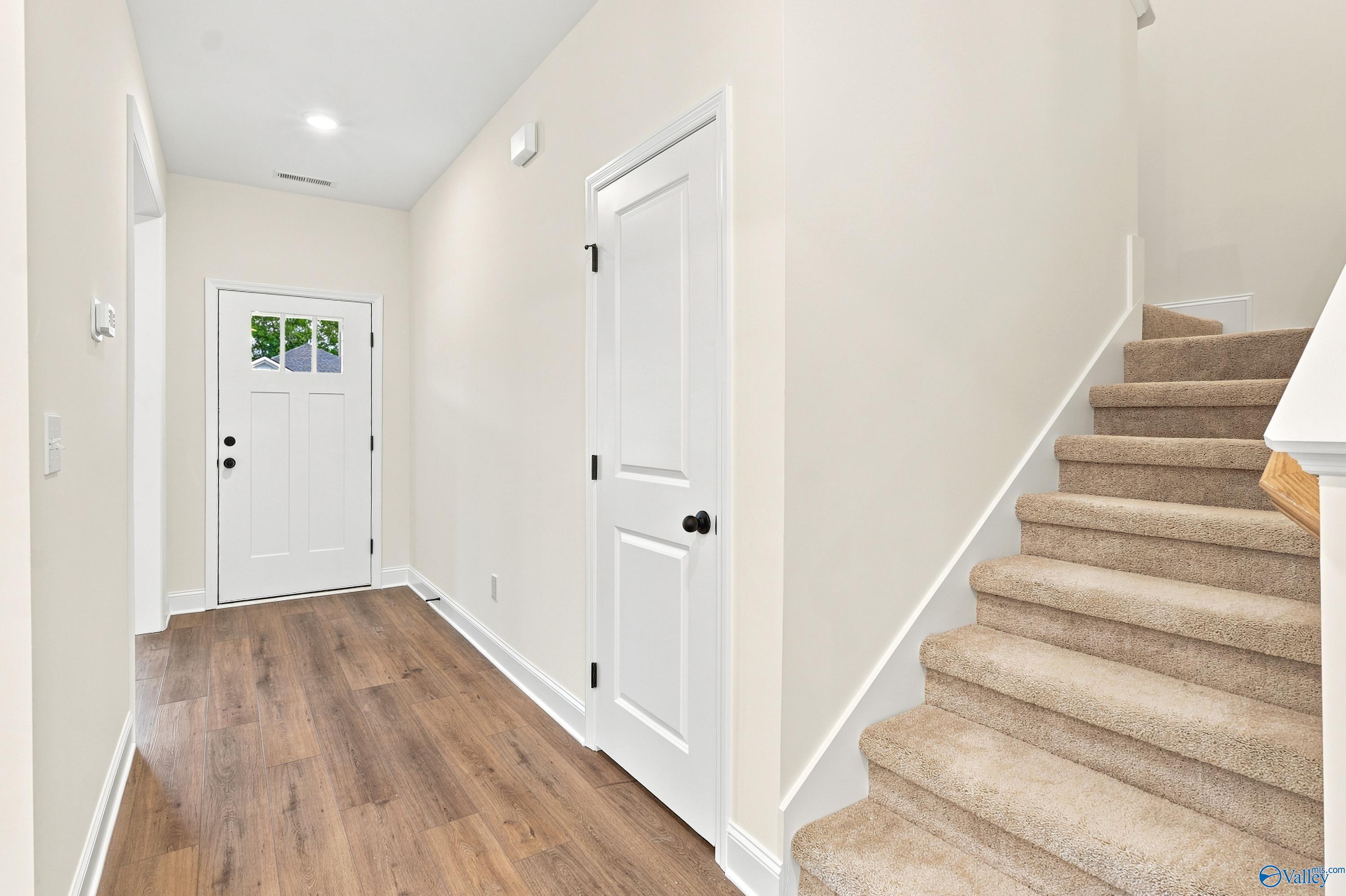 Bright foyer with wood-look flooring, white front door, and carpeted stairs in The Shelby D home at Noble Ridge, Cullman.