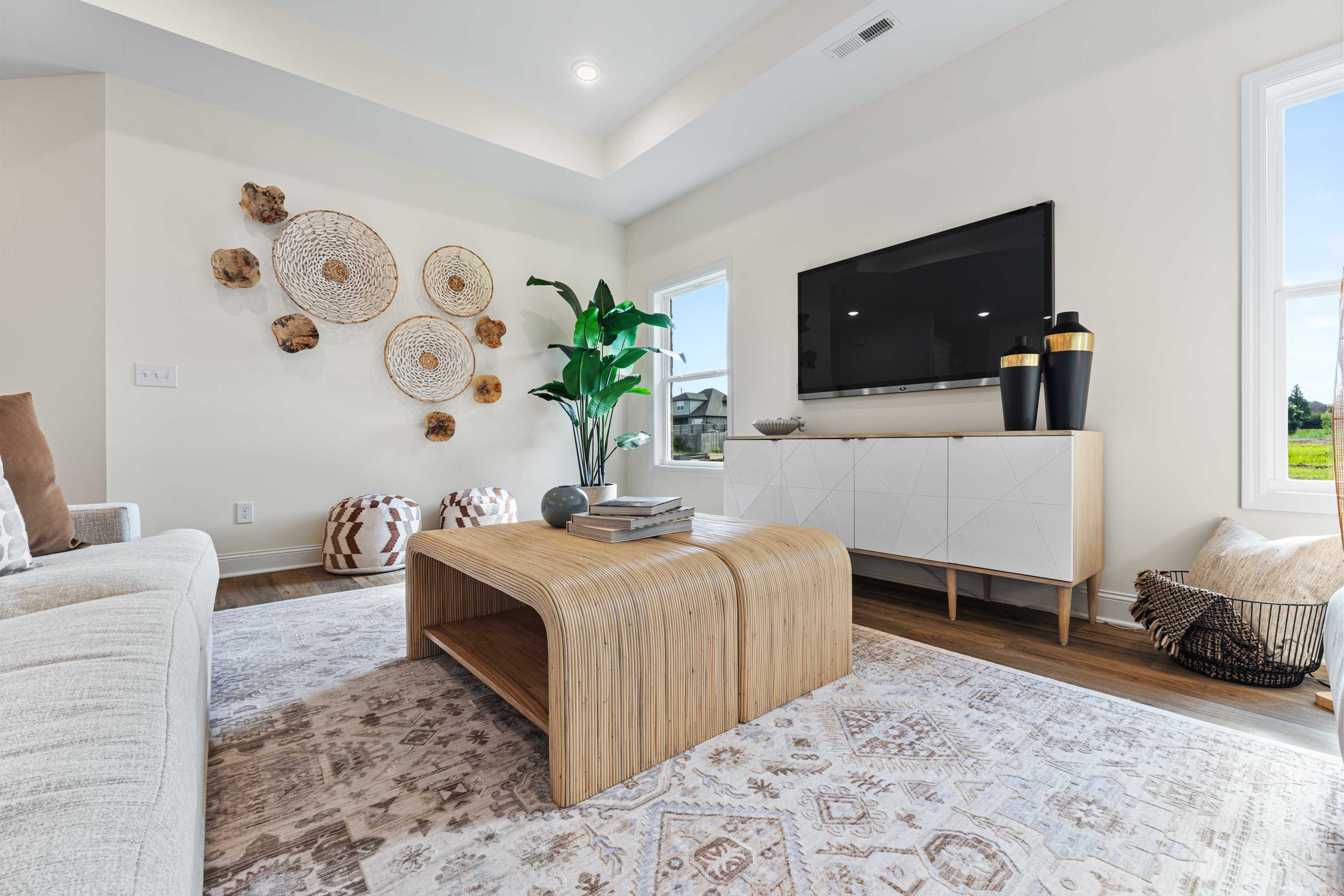 Cozy Lynn Meadows living room in Meridianville AL with woven wall decor, potted plant, beige sofa, and modern TV by Davidson Homes