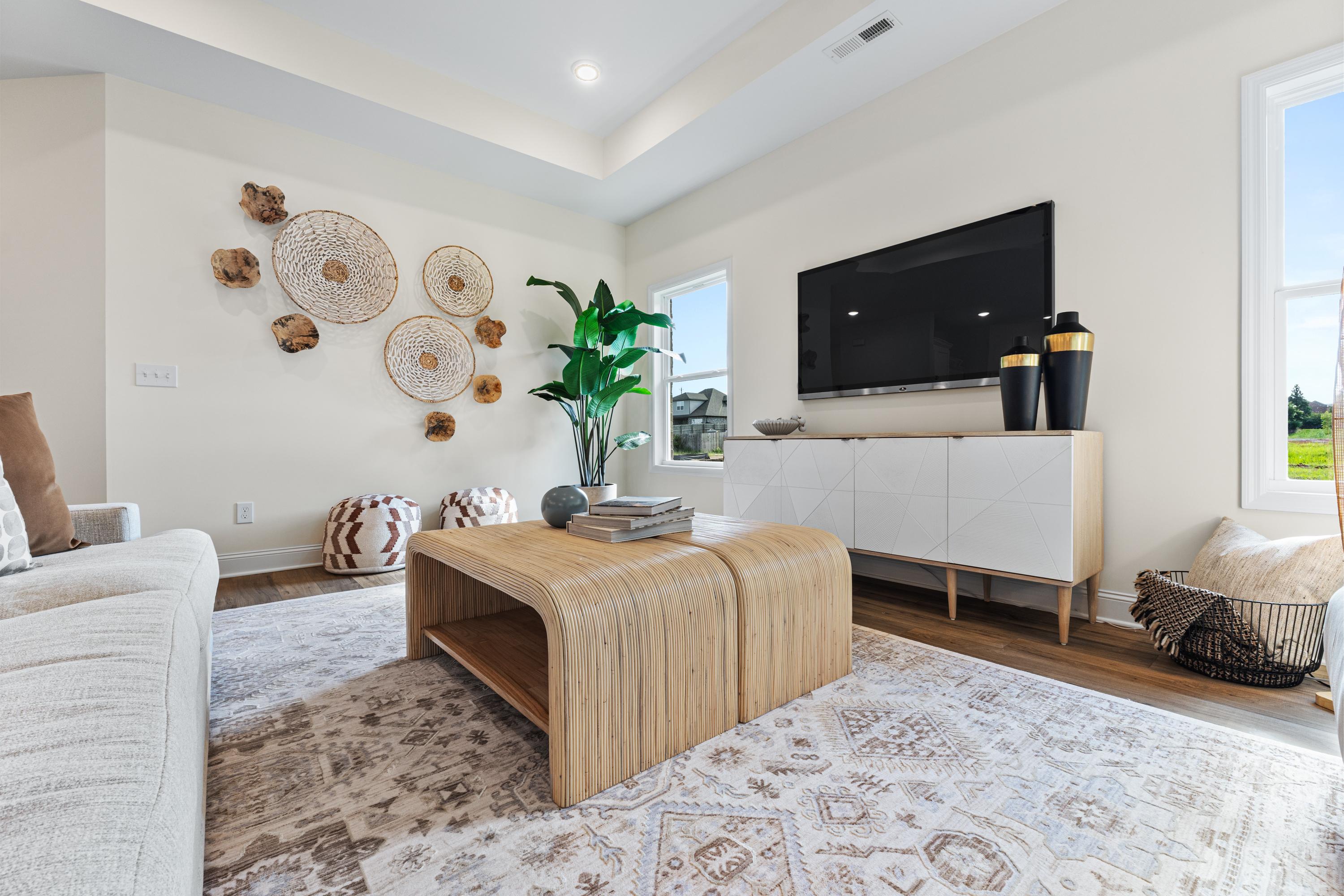 Cozy Lynn Meadows living room in Meridianville AL with woven wall decor, potted plant, beige sofa, and modern TV by Davidson Homes