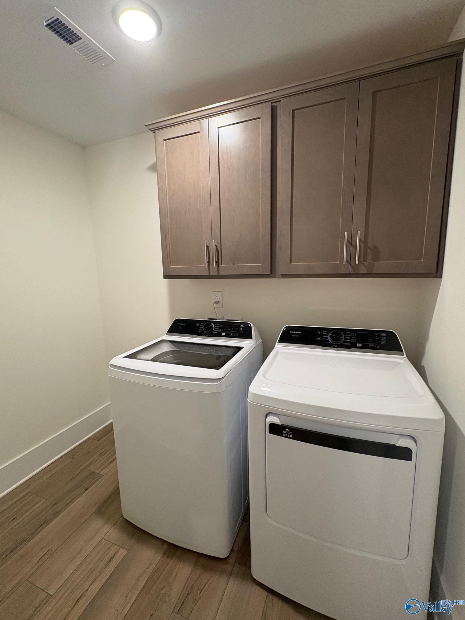 Modern laundry room featuring white front-load washer and dryer with upper wood cabinets in Davidson Homes The Daphne C, Arab, Alabama
