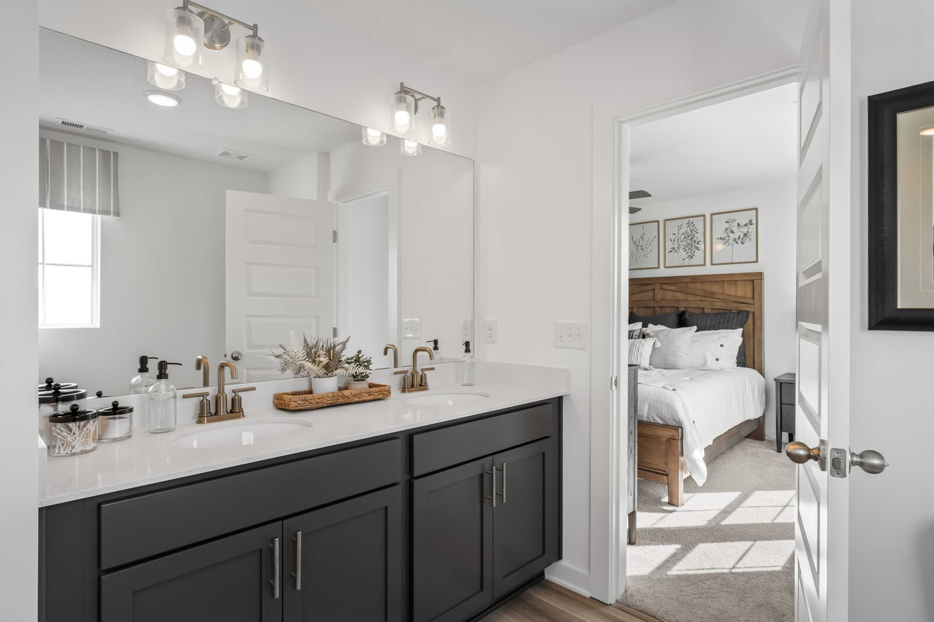 Spacious primary bathroom featuring dark shaker vanity, gold faucets, and open door to cozy bedroom at The Towns at Red River in Gallatin TN