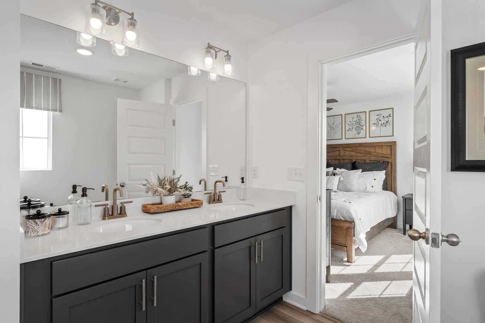 Spacious primary bathroom featuring dark shaker vanity, gold faucets, and open door to cozy bedroom at The Towns at Red River in Gallatin TN