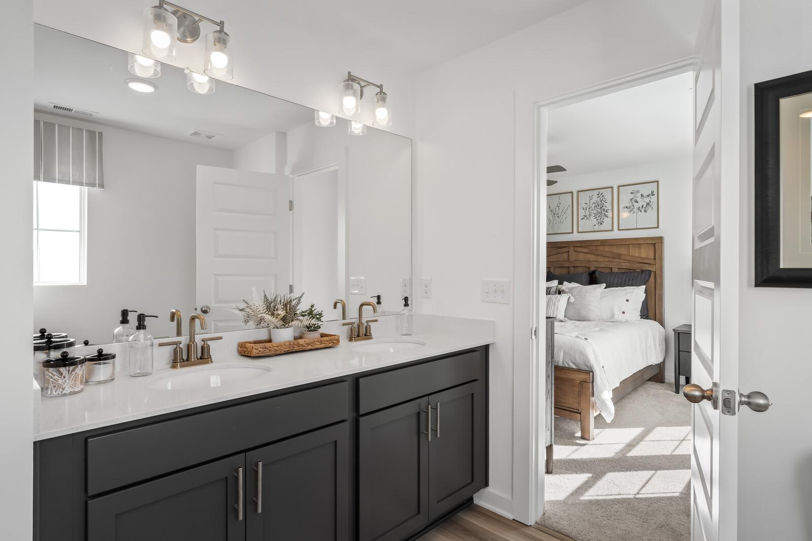 Spacious primary bathroom featuring dark shaker vanity, gold faucets, and open door to cozy bedroom at The Towns at Red River in Gallatin TN
