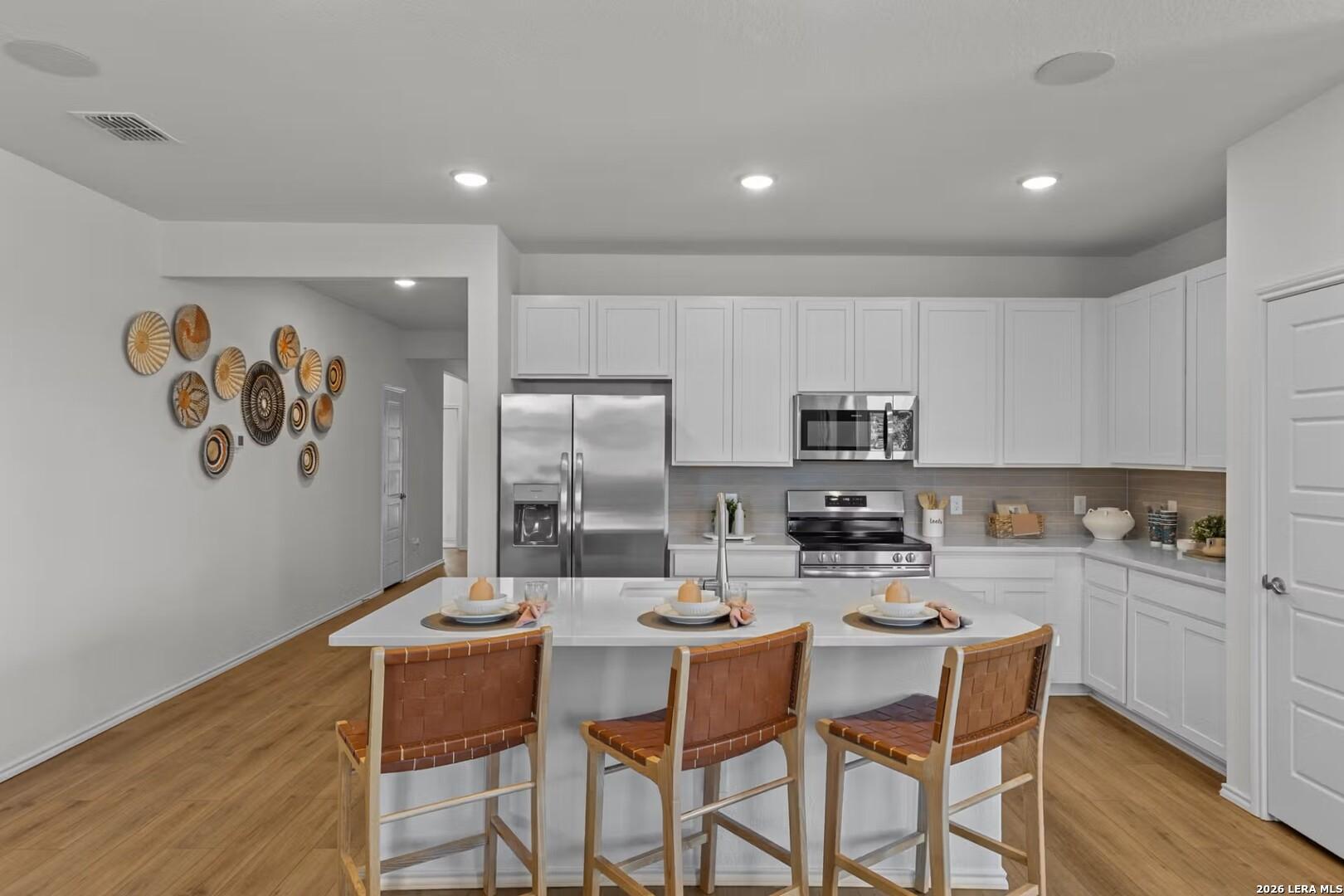 Modern white kitchen with stainless steel appliances, quartz island, rattan bar stools, and woven wall decor in The Daphne K, Bricewood, San Antonio