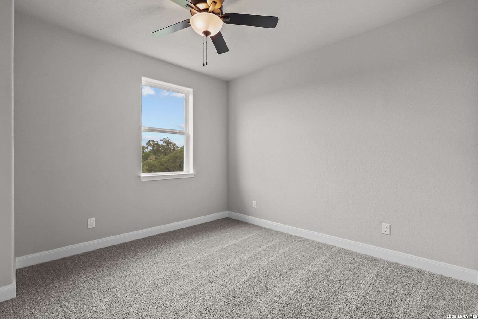 Bright bedroom with ceiling fan, large window overlooking trees, gray walls and carpet in Davidson Homes The Belmont H, Castroville, Texas