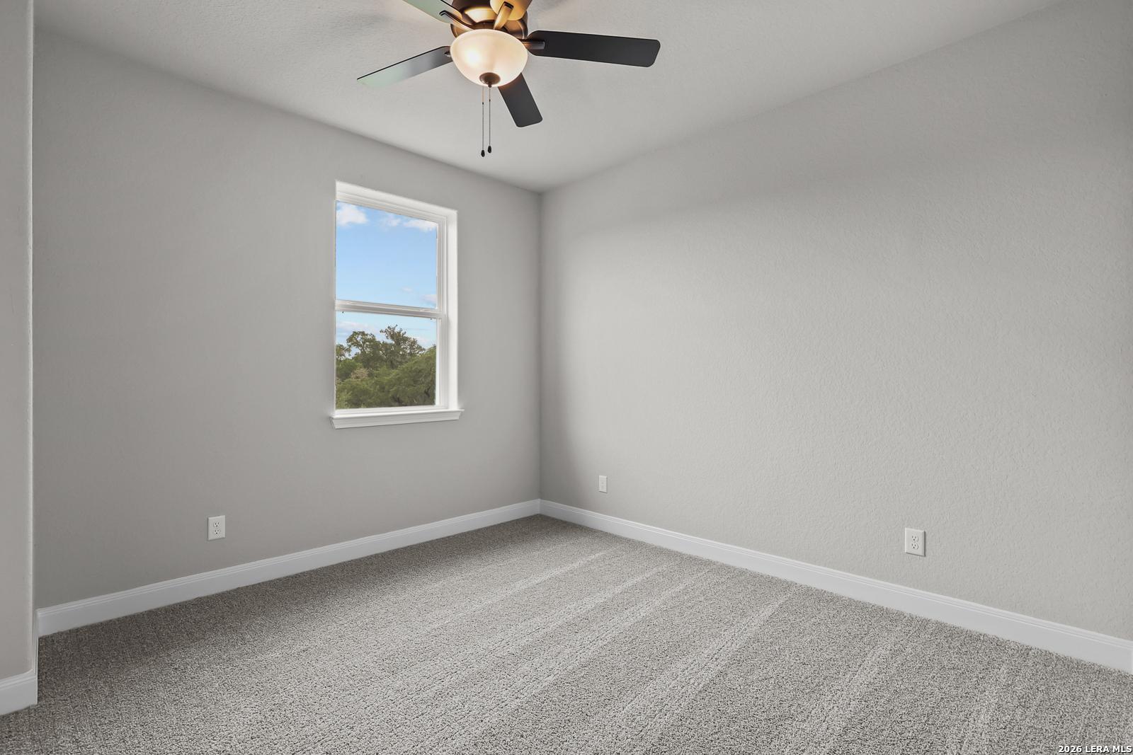 Bright bedroom with ceiling fan, large window overlooking trees, gray walls and carpet in Davidson Homes The Belmont H, Castroville, Texas