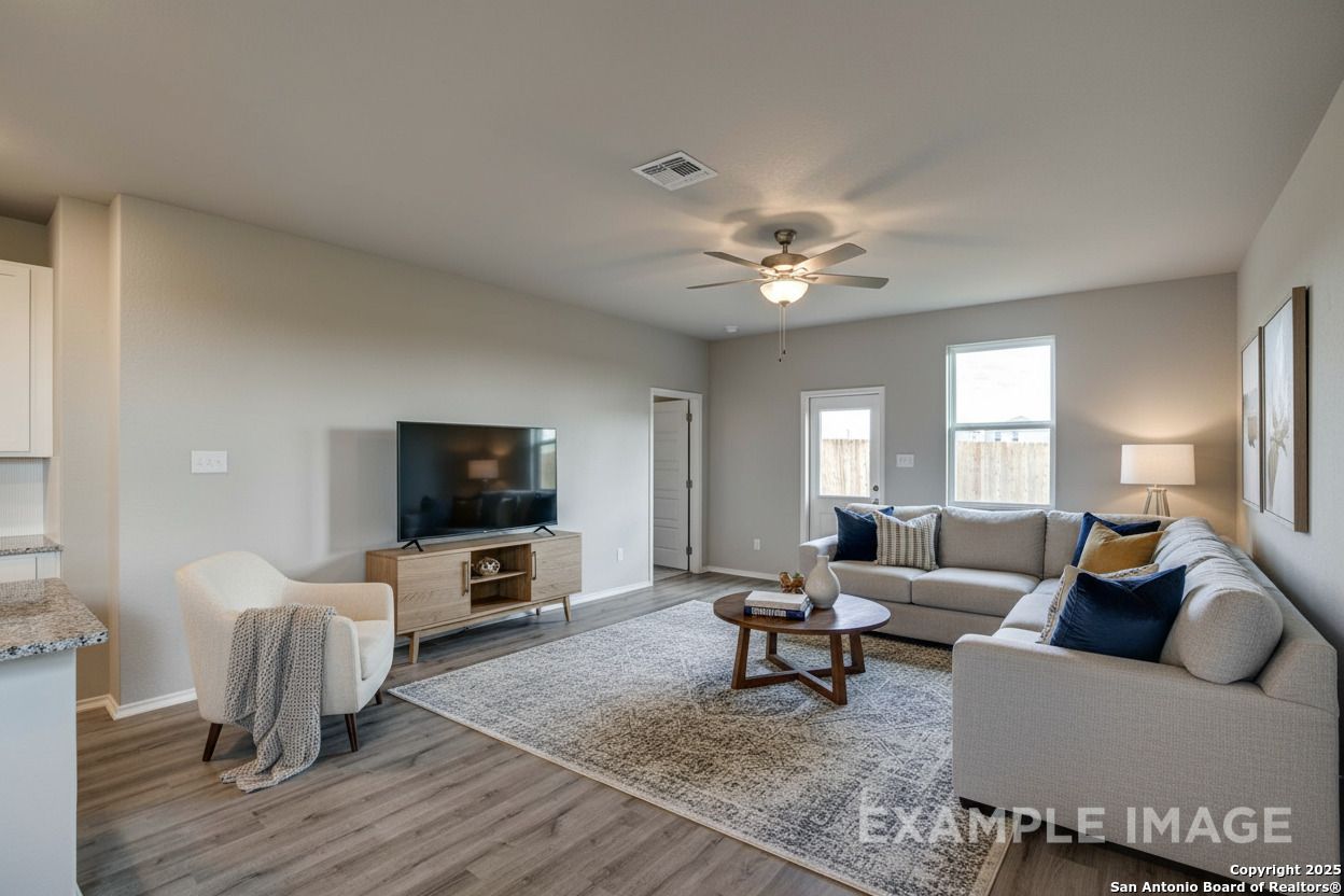 Spacious living room with neutral walls, L-shaped sofa, wall TV, ceiling fan, and open kitchen in Davidson Homes The Blanco C, San Antonio