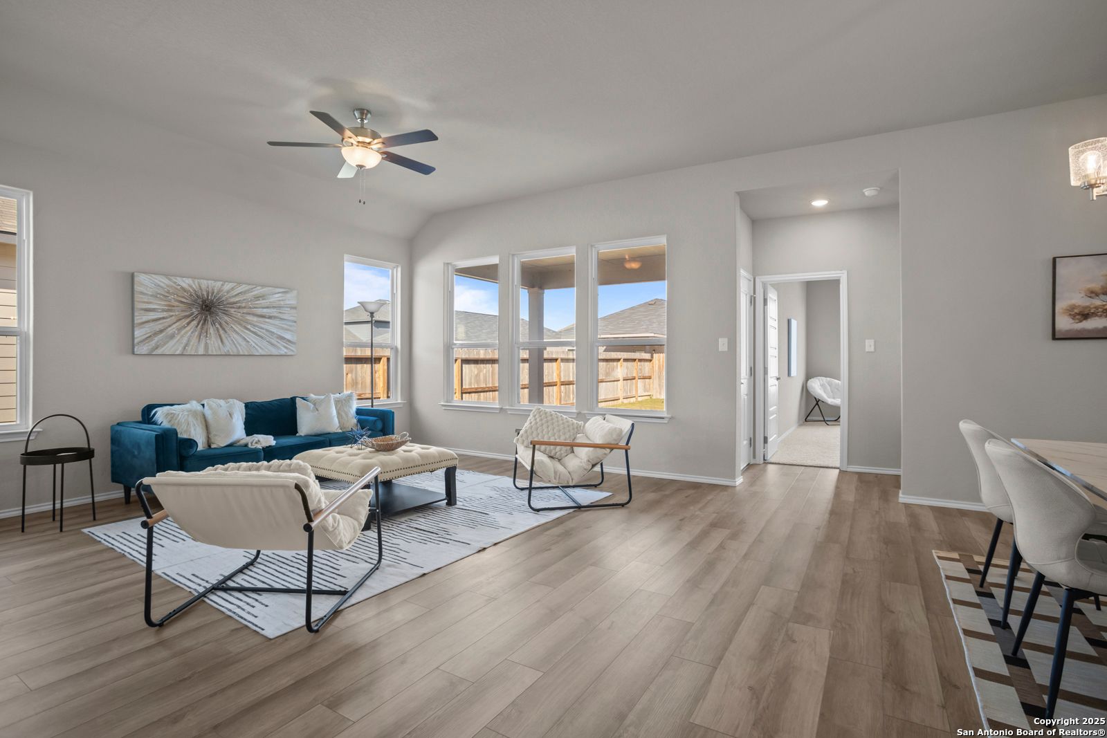 Modern open-concept living room with blue velvet sofa, ceiling fan, large windows, and dining area in The Collin B home, Seguin, Texas