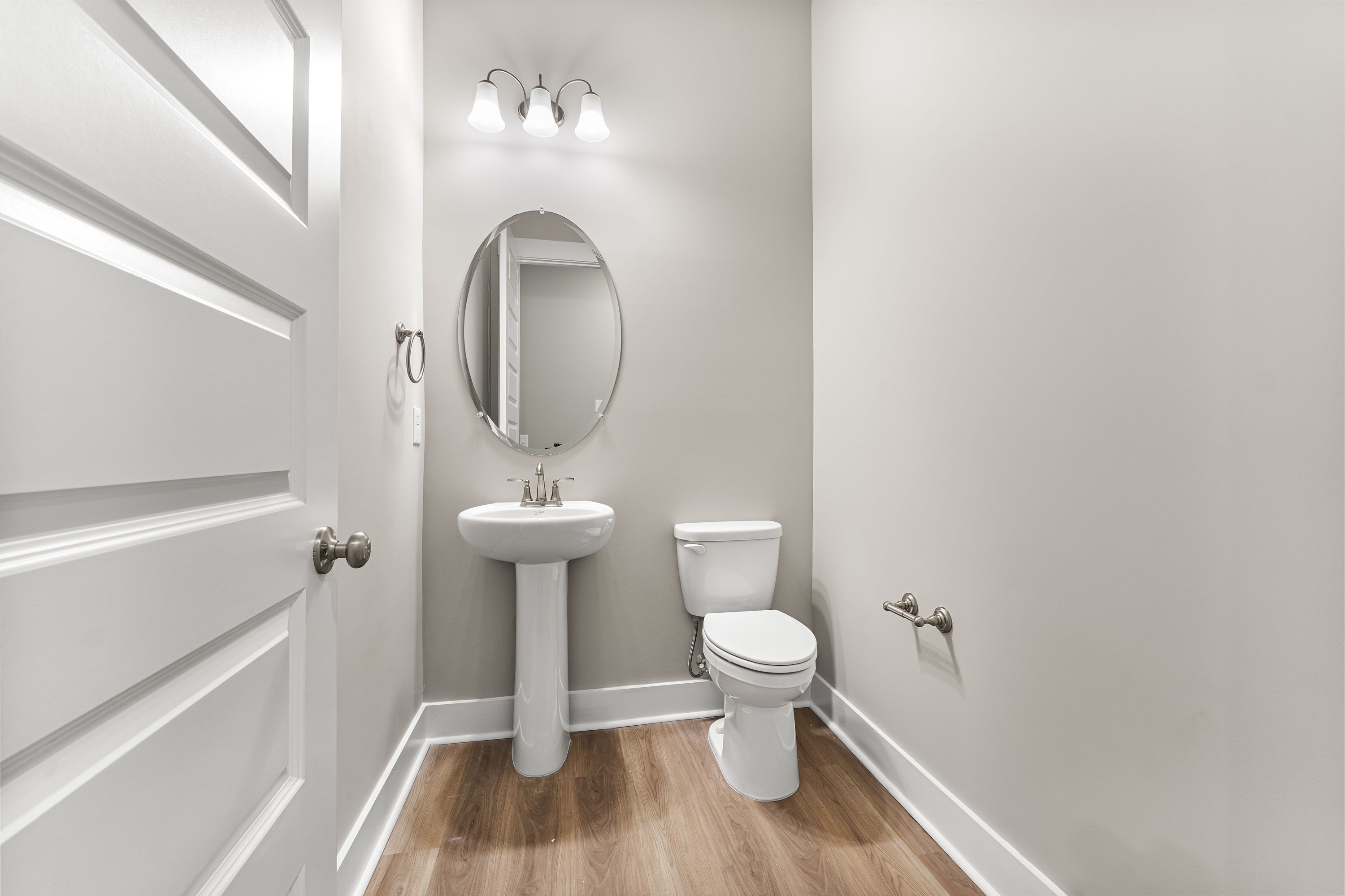 Compact powder room in The Oxford A showcasing pedestal sink, round mirror, and neutral walls with wood flooring