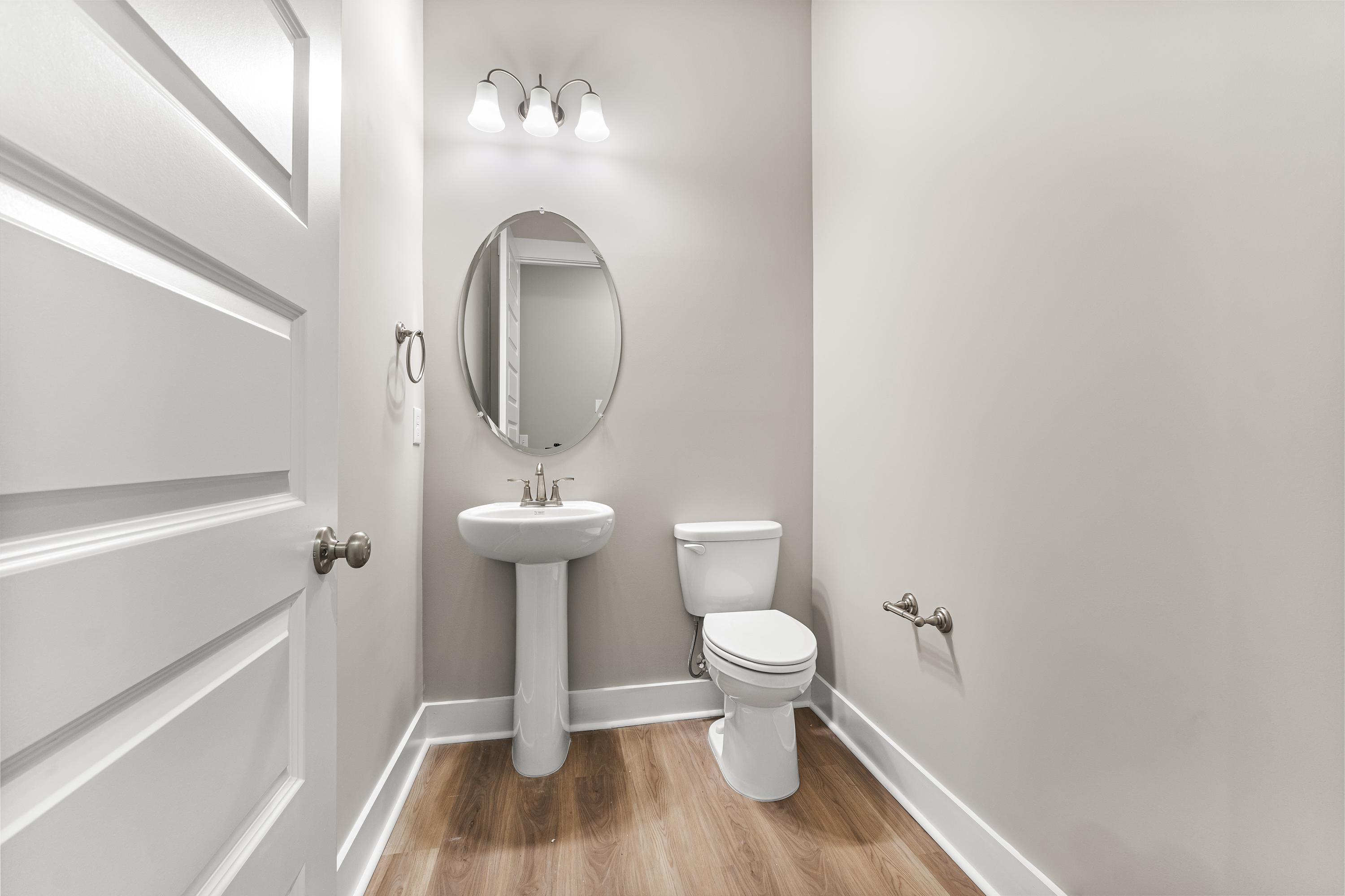 Compact powder room in The Oxford A showcasing pedestal sink, round mirror, and neutral walls with wood flooring