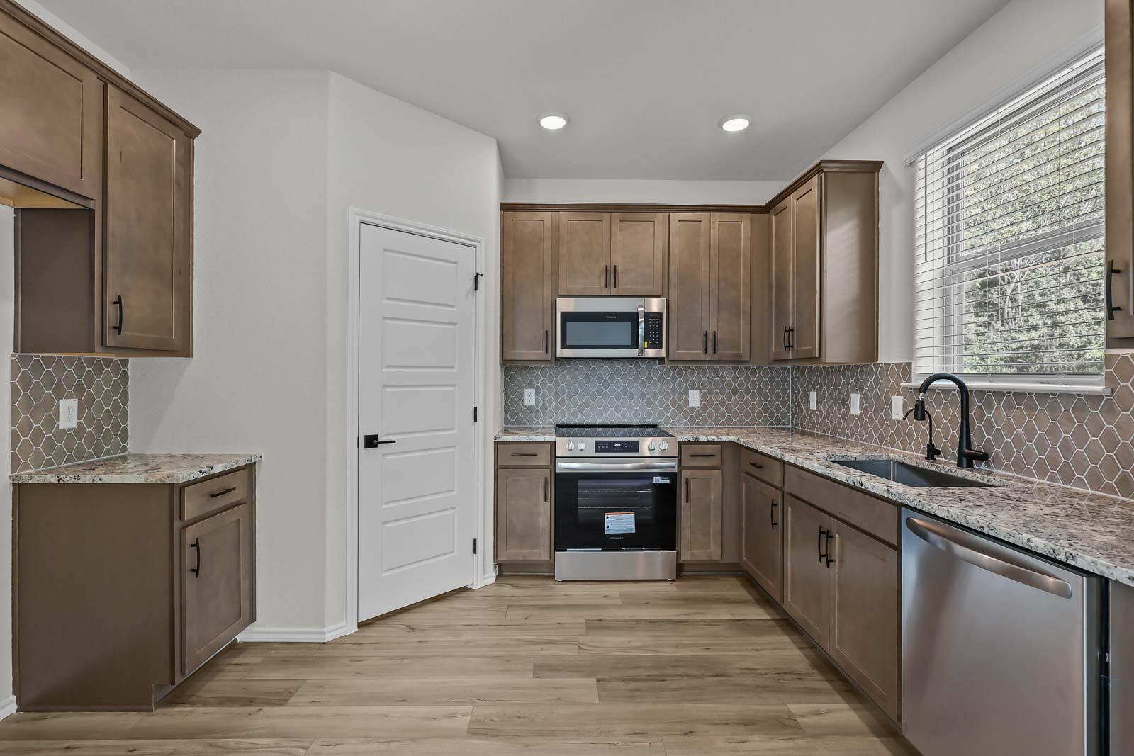 Modern kitchen in The Florence B featuring brown cabinetry, granite countertops, tile backsplash, and stainless appliances.