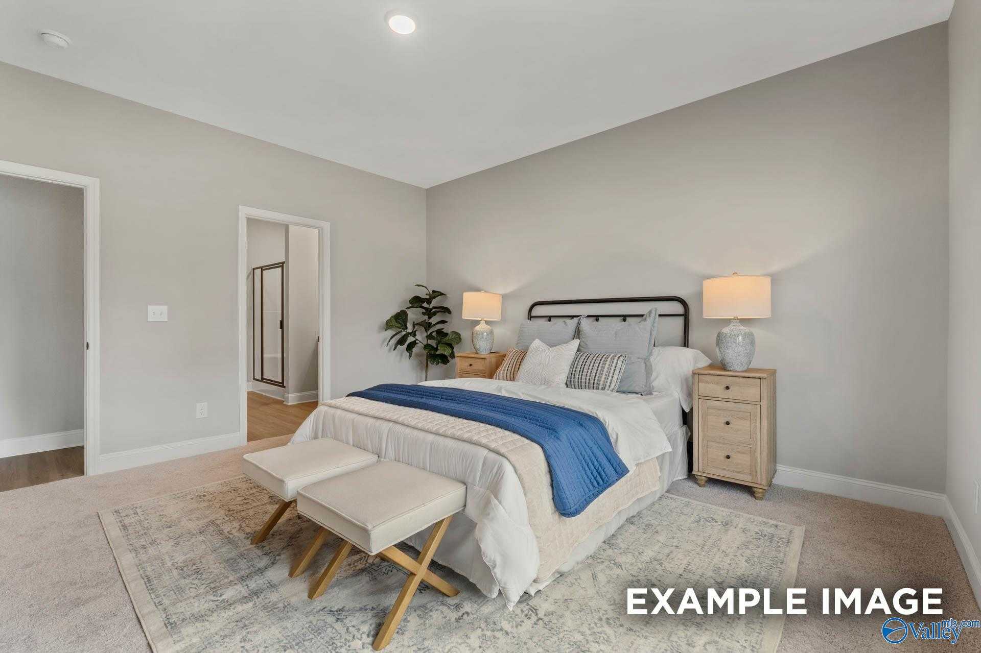 Cozy master bedroom with blue duvet, wooden nightstands, lamps, and ottomans in Davidson Homes The Daphne C, Owens Cross Roads, AL