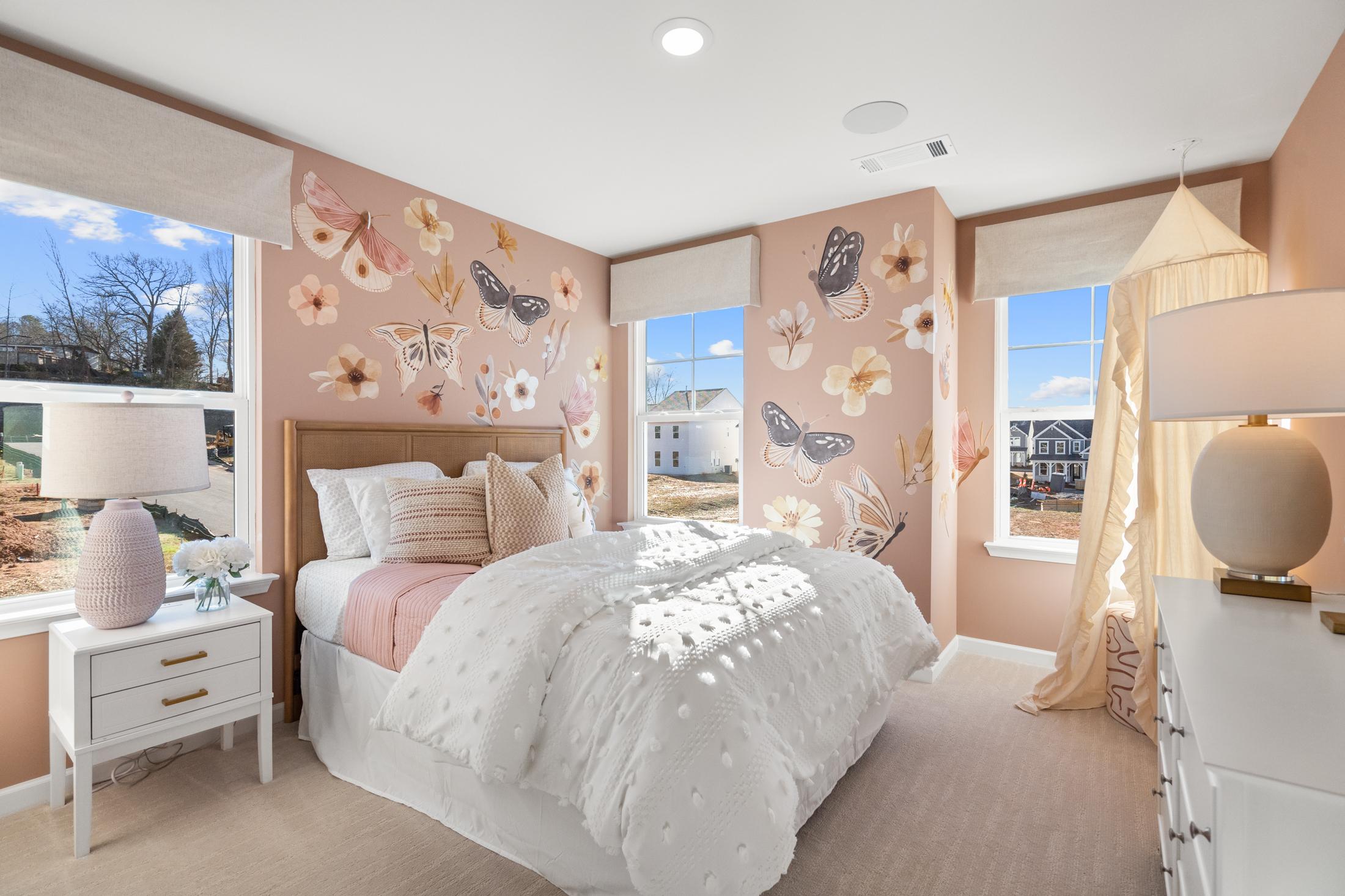 Charming girl's bedroom in The Marion A with pink floral butterfly wallpaper, tufted bed, white dresser, and sunny windows