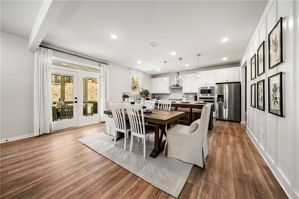 Spacious dining area with wooden farmhouse table, white chairs, and adjacent white kitchen with stainless appliances in The Willow B home, Riverwood, Dallas, Georgia