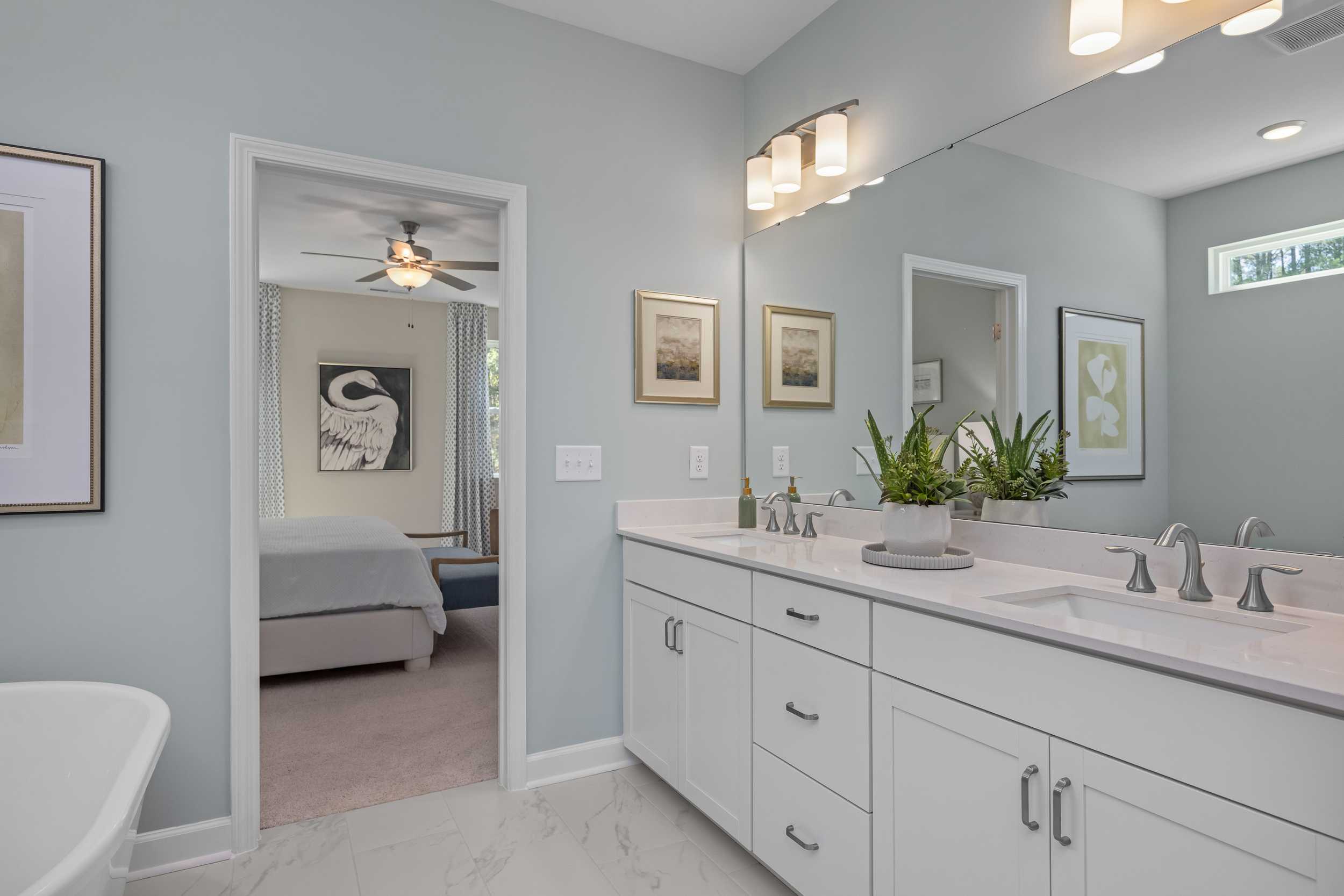 Spacious master bathroom in The Hickory II E with double vanity, light blue walls, artwork, and bedroom view through open door