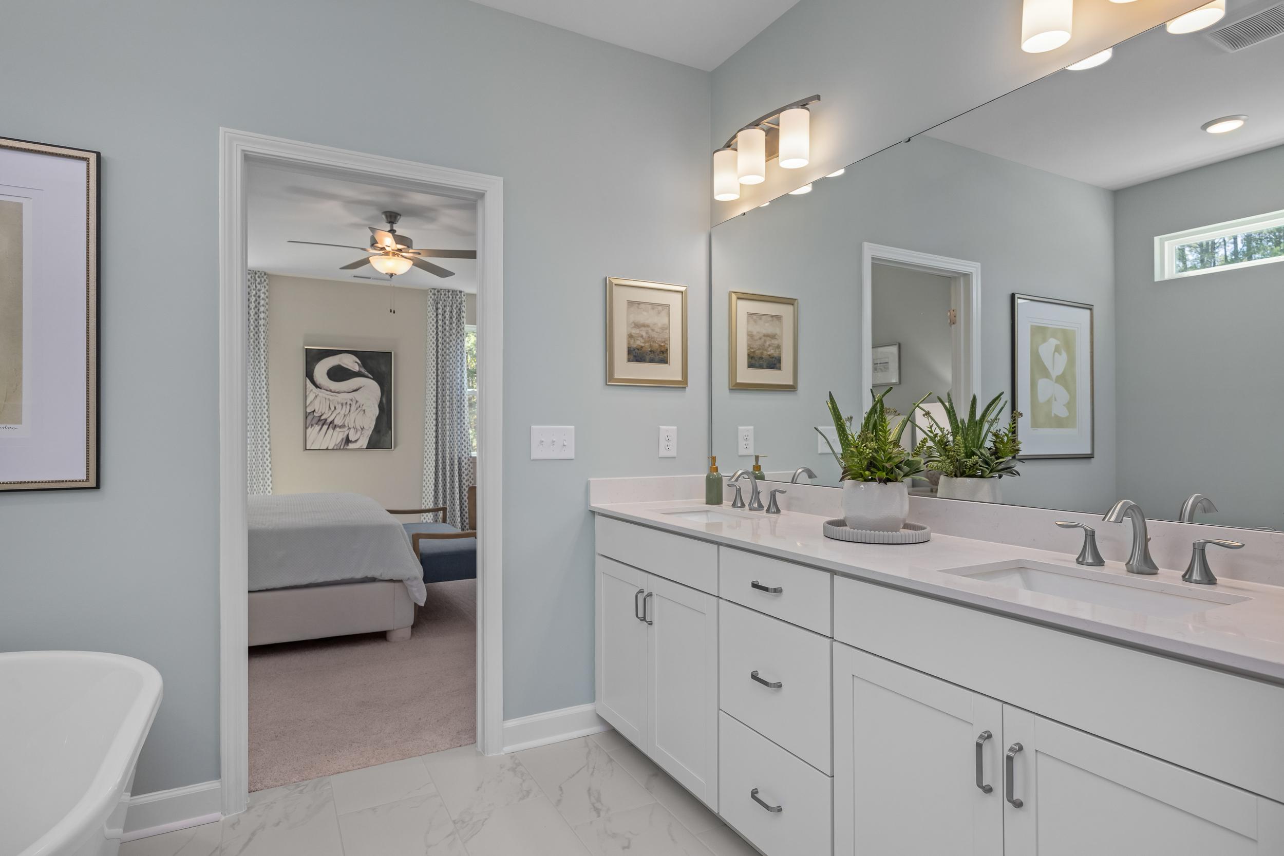 Spacious master bathroom in The Hickory II with double vanity, soft blue walls, mirrors, artwork, and bedroom view