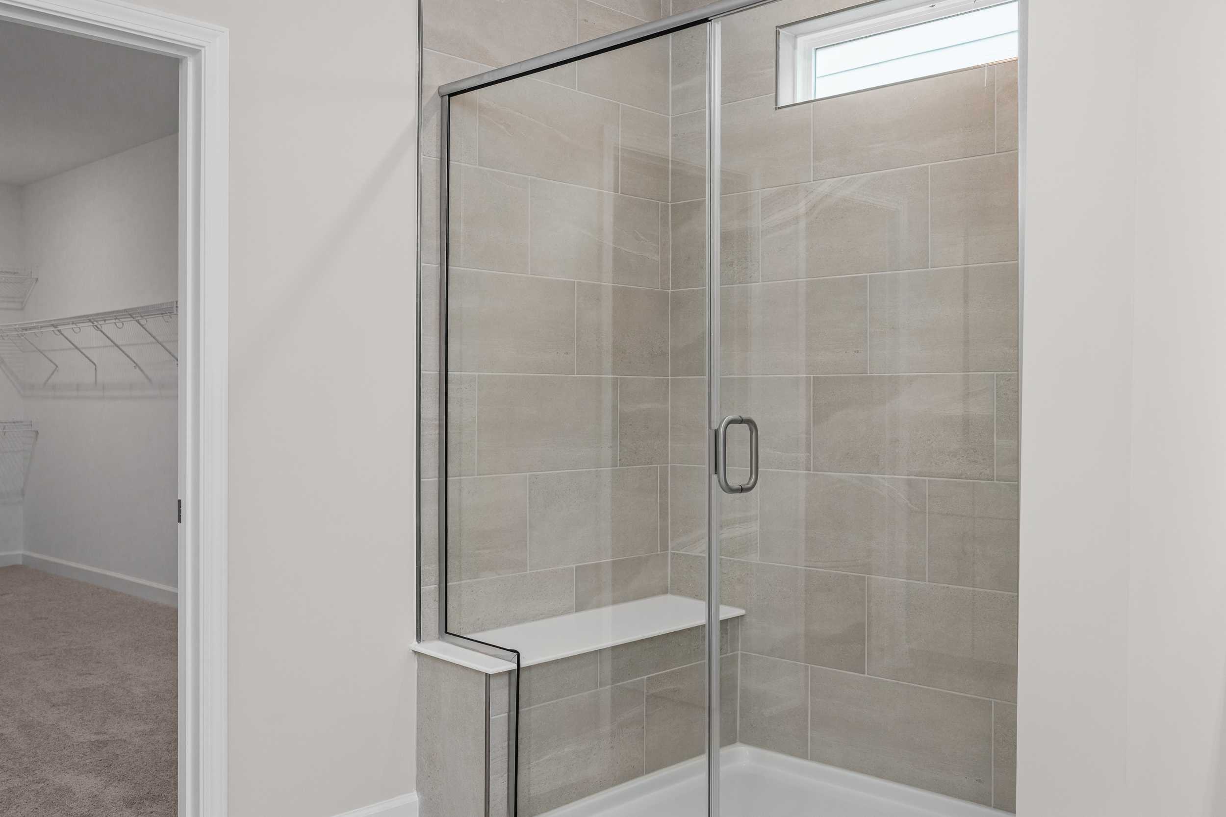 Spacious walk-in shower in The Aspen master suite with frameless glass doors, beige tile, built-in bench, and adjacent closet