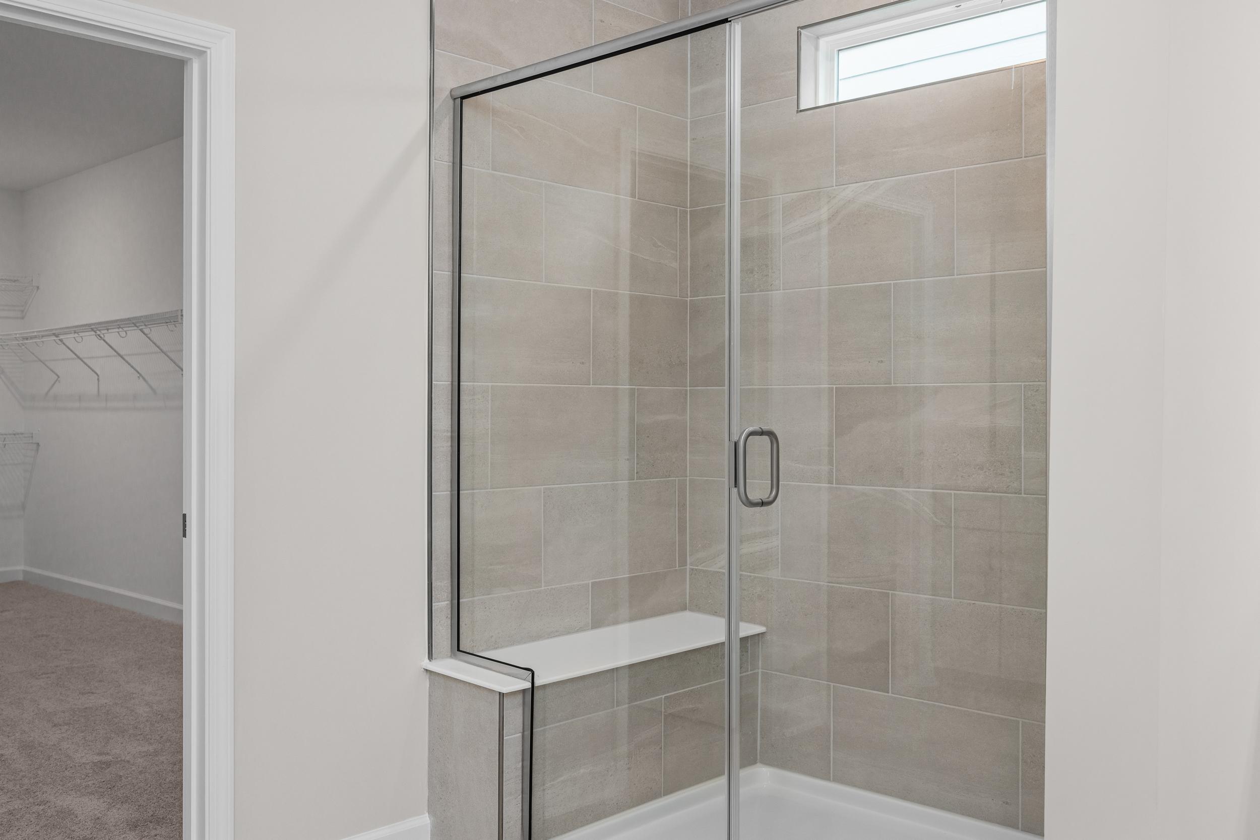 Spacious walk-in shower in The Aspen master suite with frameless glass doors, beige tile, built-in bench, and adjacent closet