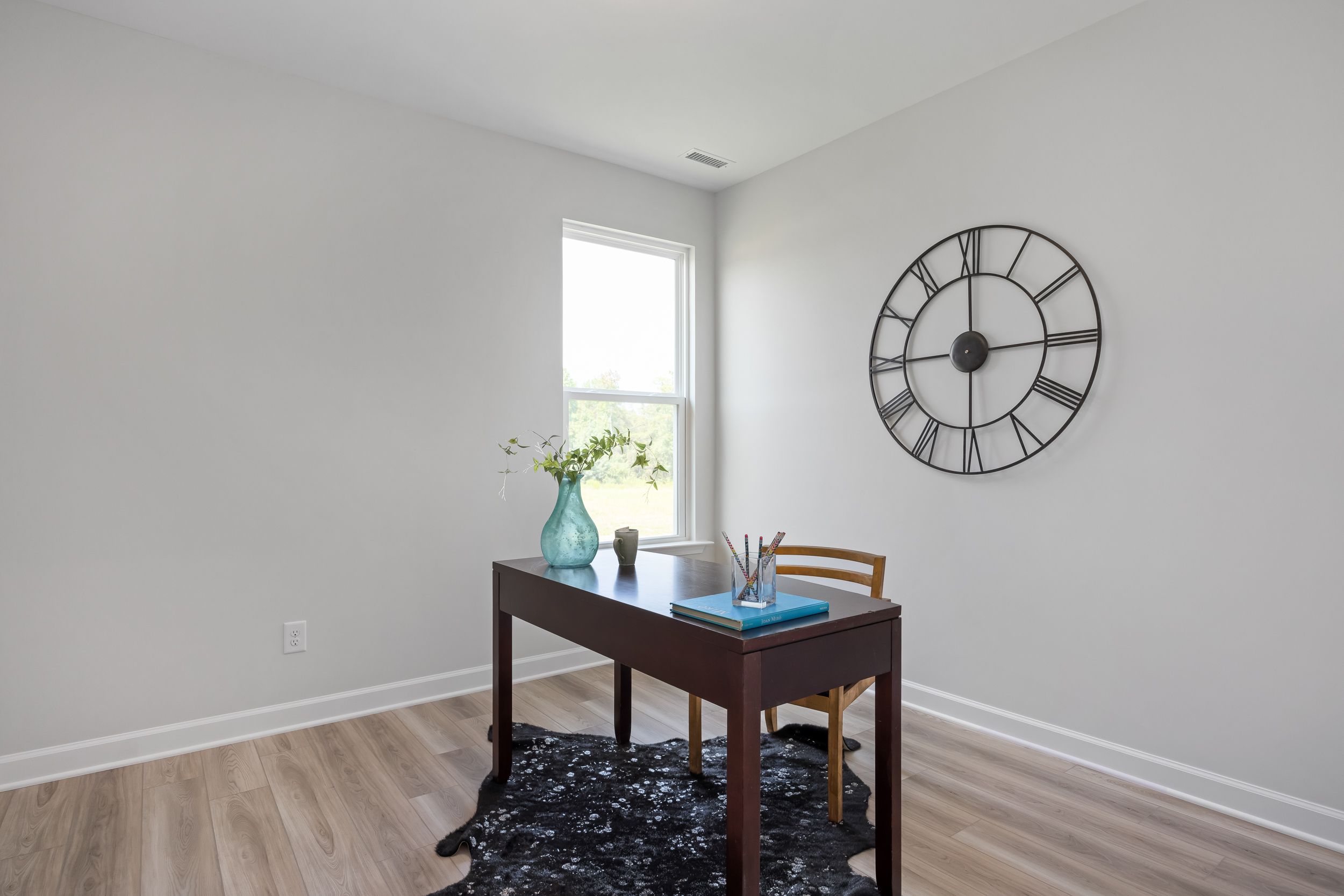 Cozy home office in The Daphne C design featuring wooden desk, blue vase of flowers, large wall clock, and natural light from window