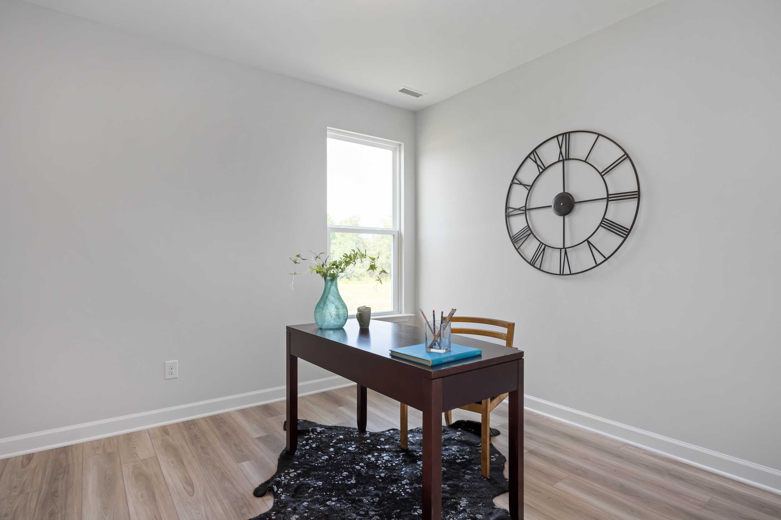 Cozy home office in The Daphne C design featuring wooden desk, blue vase of flowers, large wall clock, and natural light from window