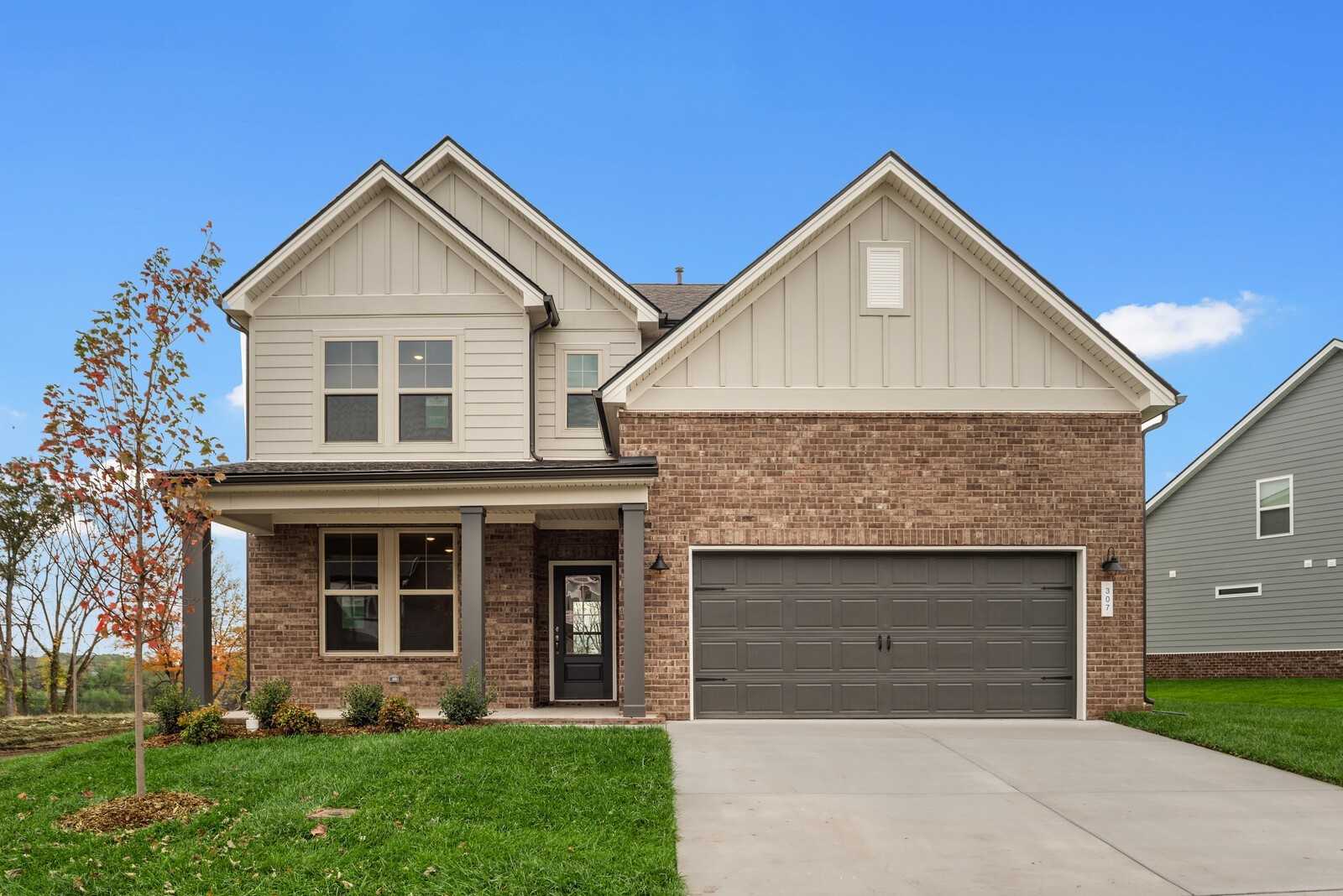 Two-story brick and siding home with covered porch, two-car garage, and autumn trees in Woods Crossing, Gallatin, Tennessee - Davidson Homes The Henry B