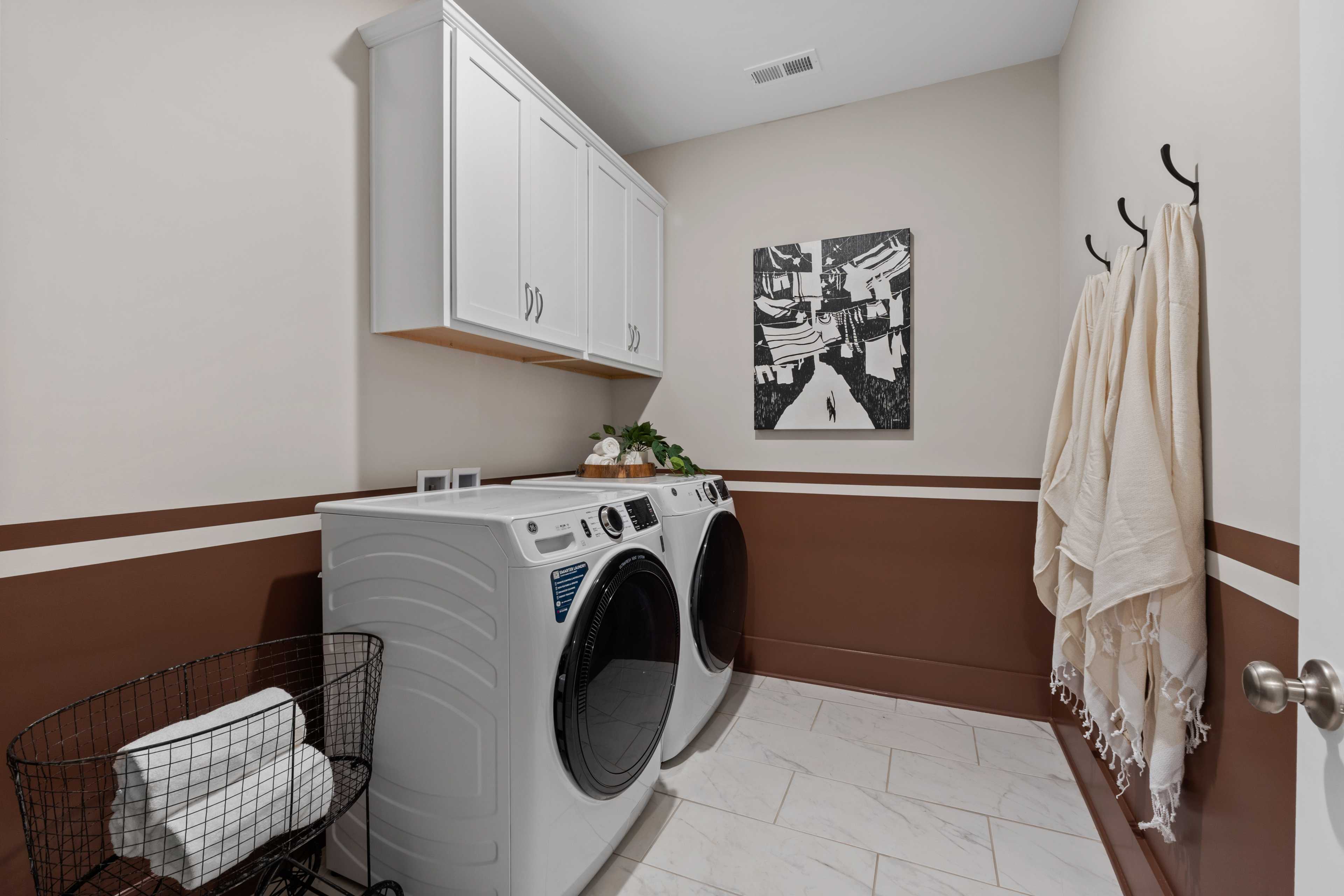 White washer dryer set in modern laundry room at Creekside Harvest AL with beige walls, upper cabinets, hanging towels, abstract art