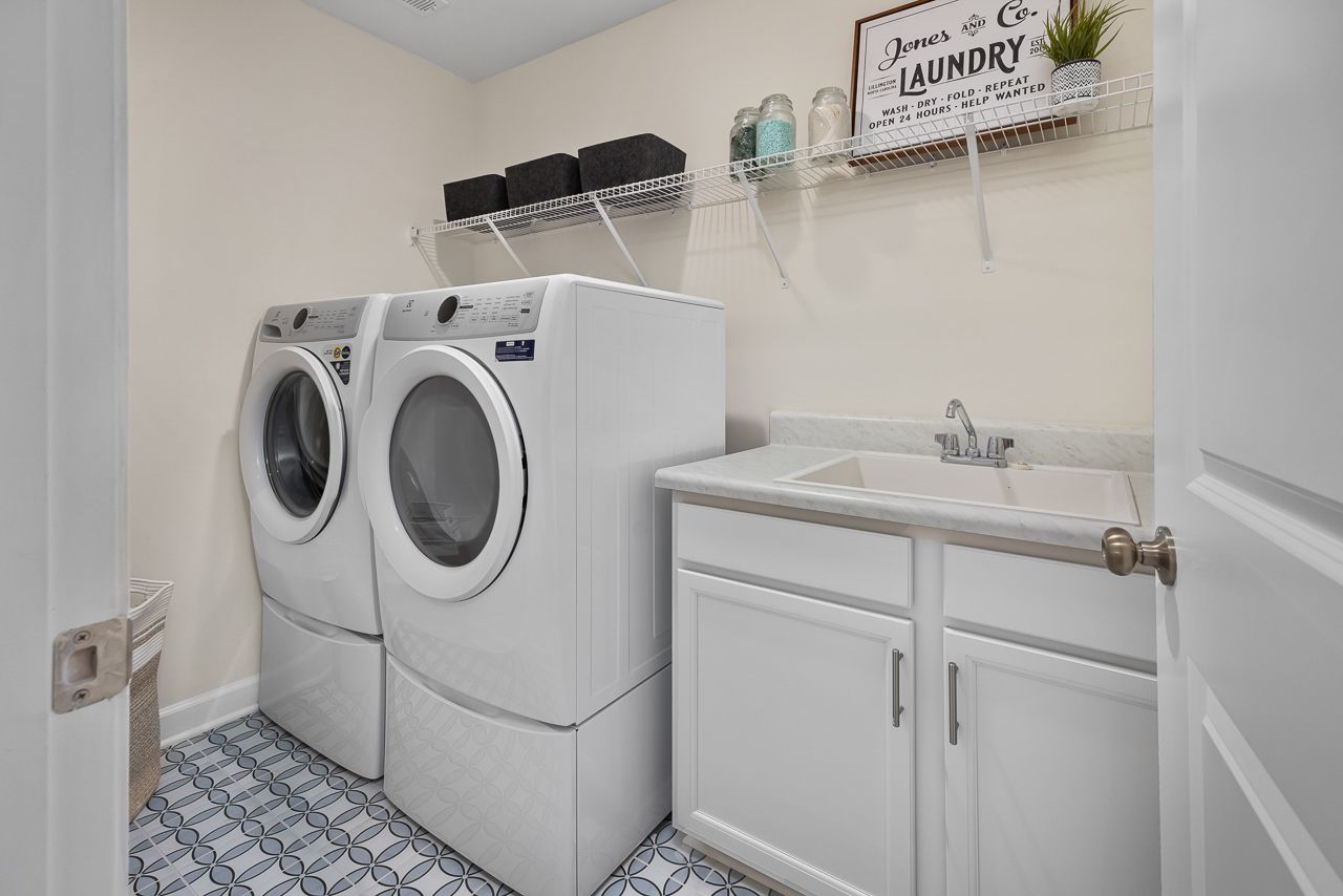 Laundry room in The Wake floor plan featuring white cabinets, utility sink, wire shelving, and modern tile flooring.