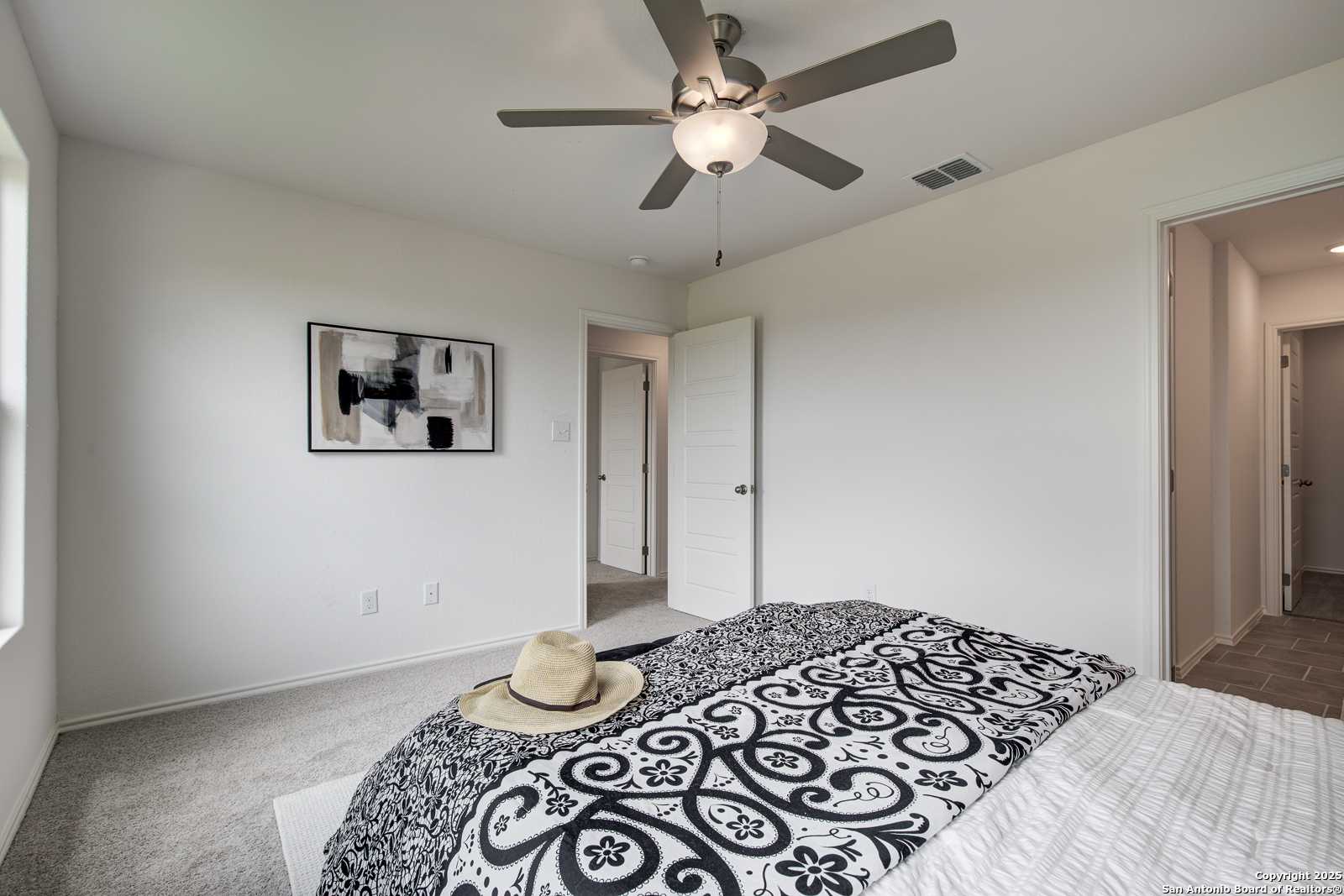Cozy bedroom with ceiling fan, patterned black-and-white bedding, abstract wall art, and adjacent bath door in Davidson Homes Trinity A, San Antonio