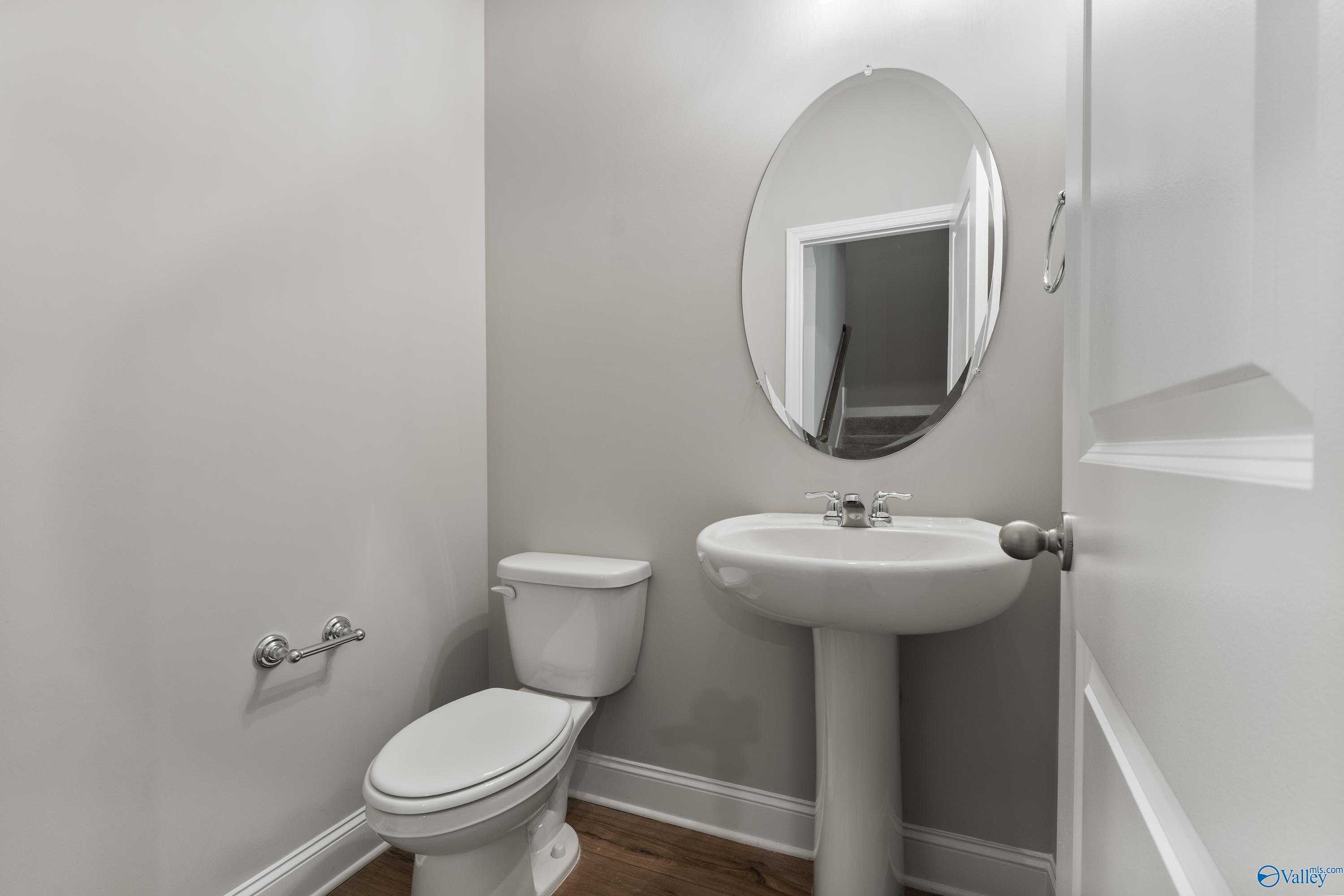 Compact powder room with white pedestal sink, oval mirror, and toilet on gray walls in Davidson Homes The Rockford, Harvest, Alabama
