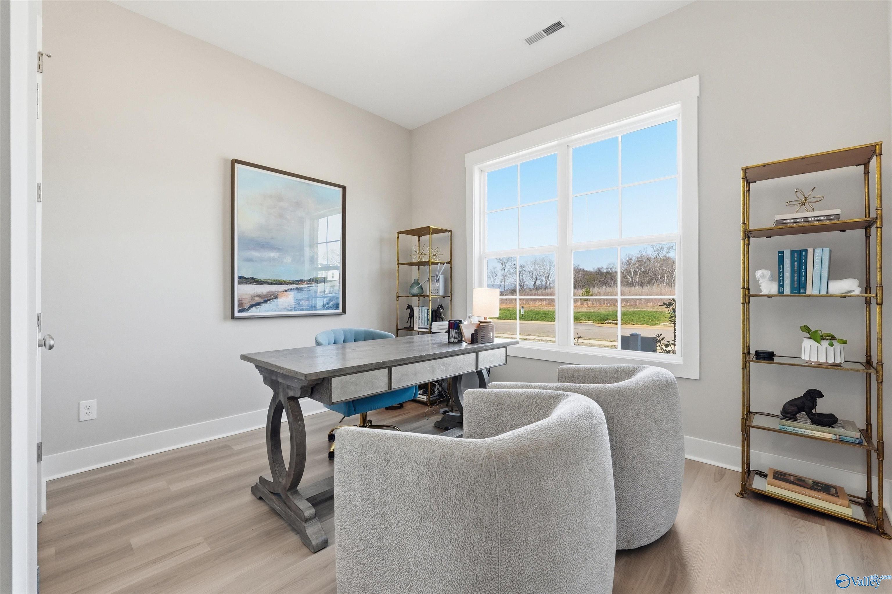 Elegant home office with wooden desk, blue chair, gray armchairs, gold bookshelf, and scenic window view in Evermore Homes Oxford B, Owens Cross Roads, Alabama
