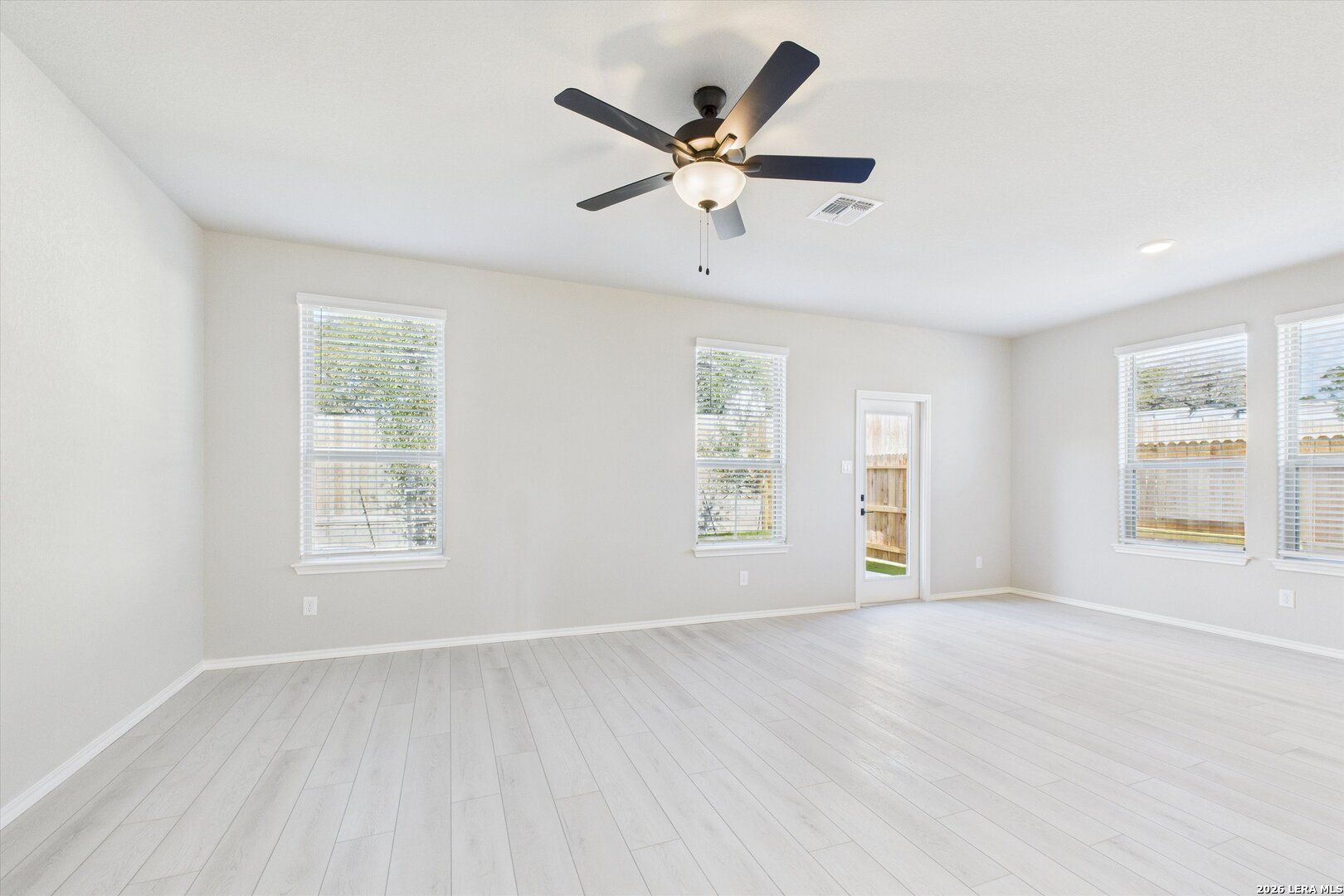 Bright empty living room with light laminate floors, beige walls, ceiling fan, and large windows in Davidson Homes The Charlotte A, San Antonio