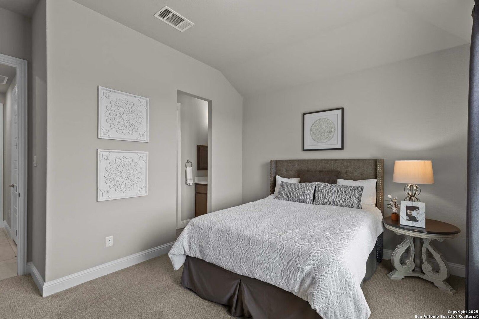Cozy secondary bedroom with queen bed, neutral gray walls, artwork, and adjacent bath in Davidson Homes The Garner B, Castroville, Texas
