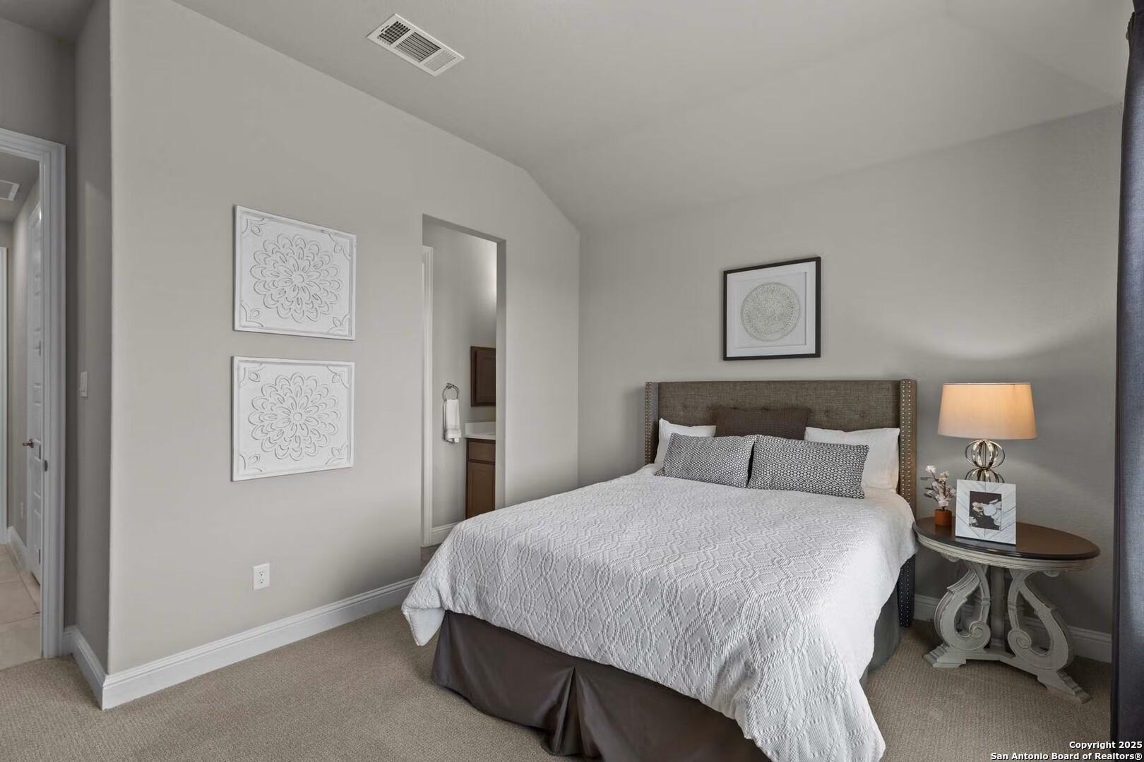 Cozy secondary bedroom with queen bed, neutral gray walls, artwork, and adjacent bath in Davidson Homes The Garner B, Castroville, Texas