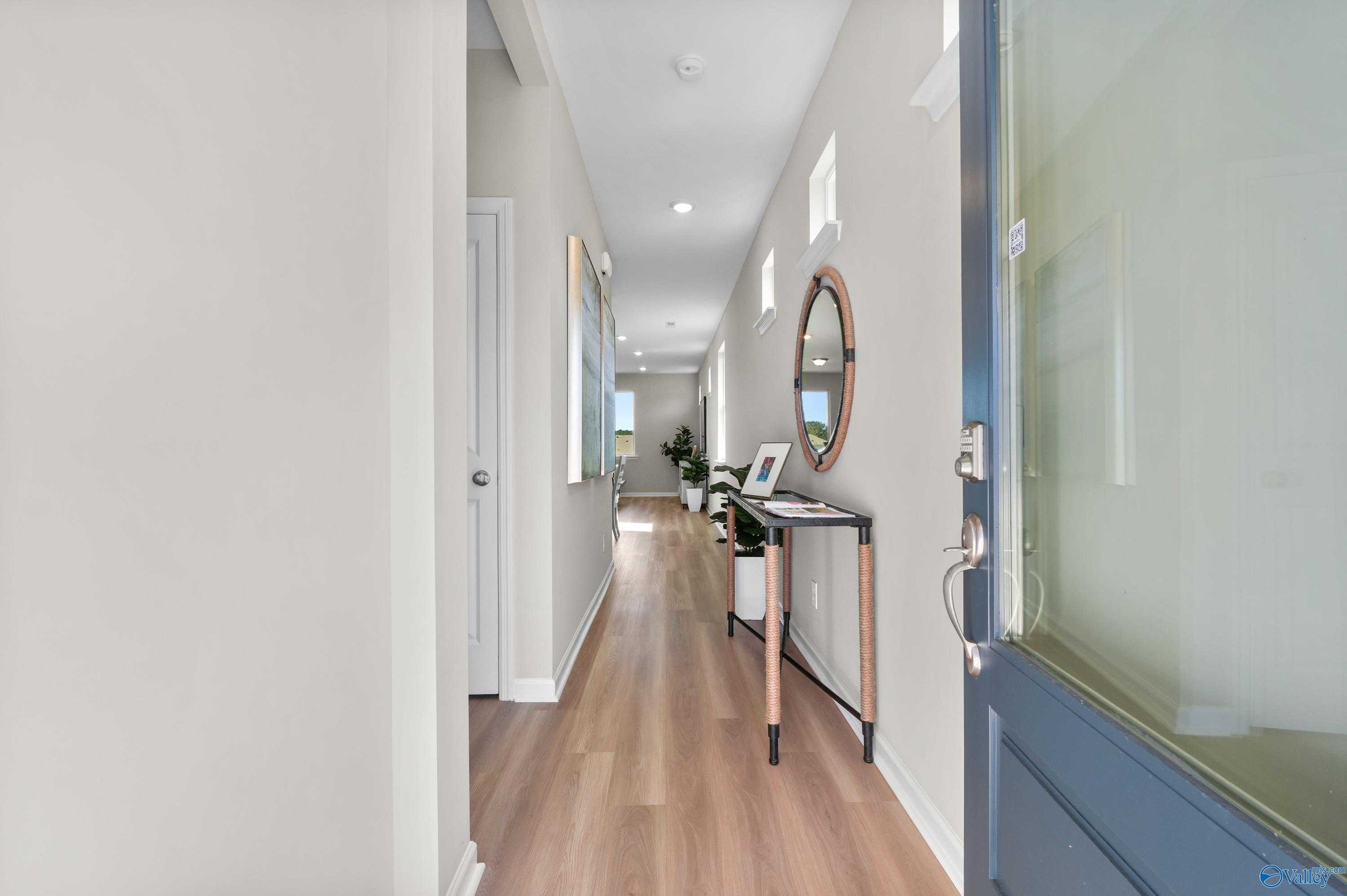 Bright entry hallway with light oak floors, round mirror, console table with plants, and blue frosted door in Evermore Homes The Malibu, Madison Alabama